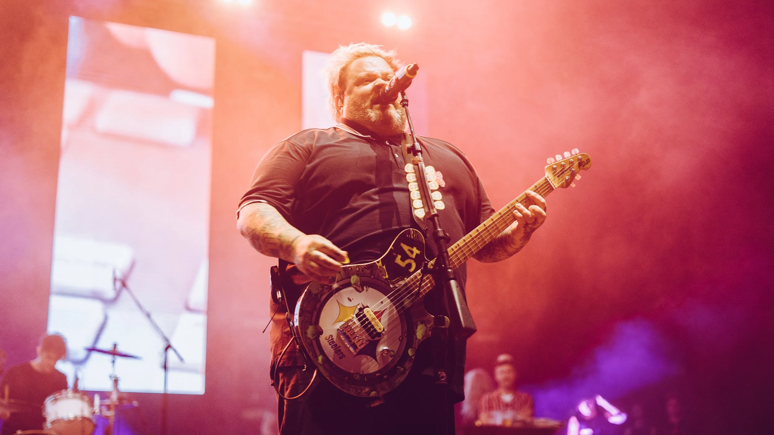 A male musician with a beard and tattoos singing into a microphone while playing a guitar on stage, with colorful lighting and digital screens in the background.