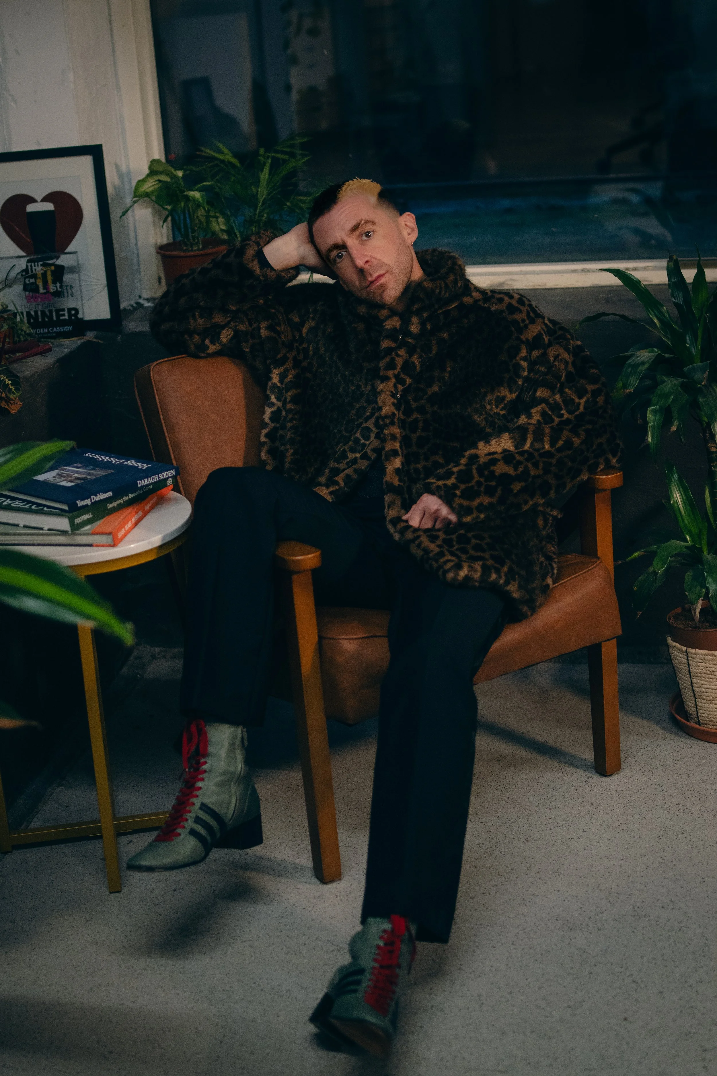 A man with short hair and a beard sitting in a brown chair, wearing a leopard-print coat, black pants, and white sneakers with red laces. He is resting his head on his left hand and looking at the camera. There are potted plants, books, and a framed 