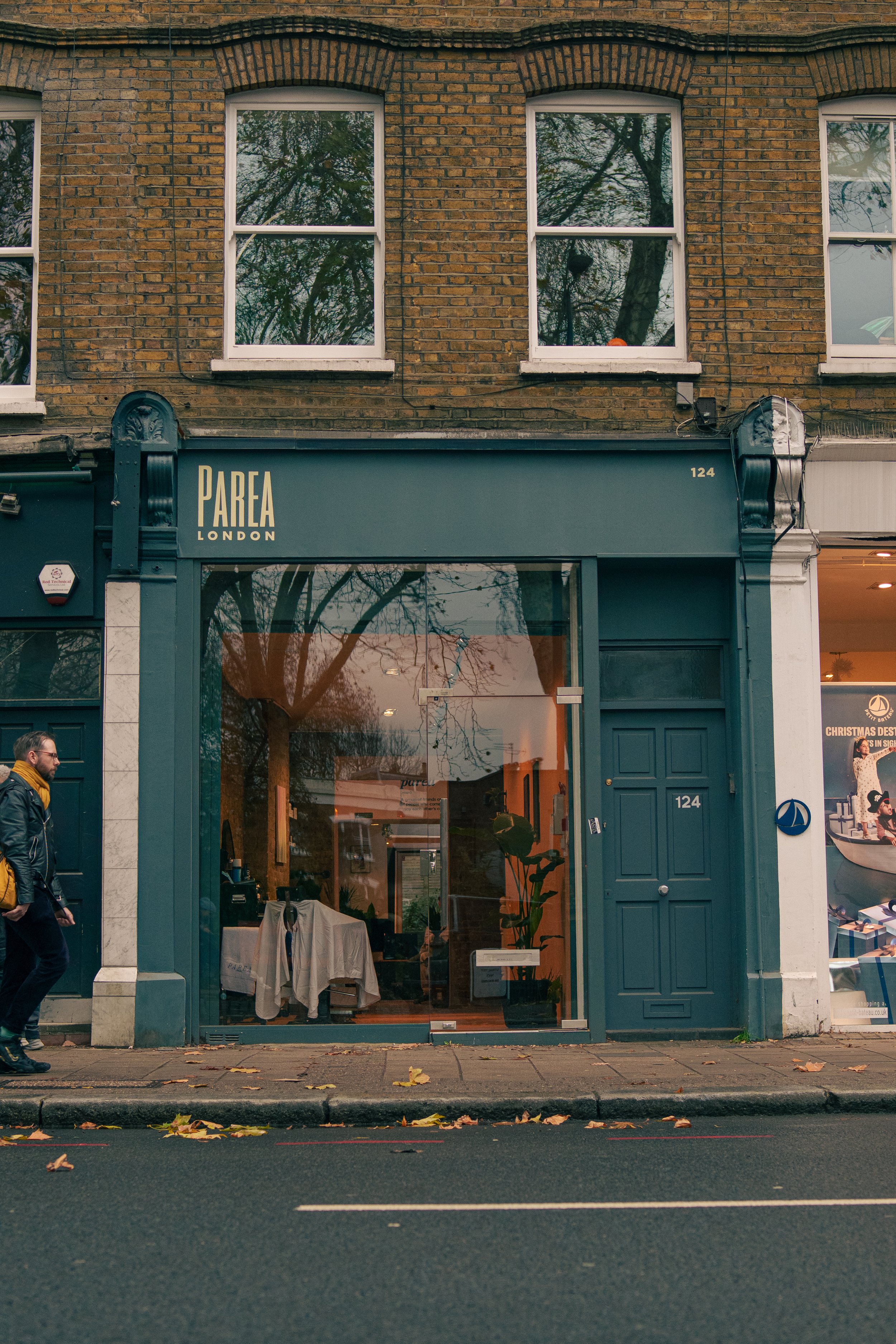  storefront with sign reading 'PAREA LONDON' at 124, a man walking on sidewalk, brick building with three upper windows, reflected trees in the storefront glass, some leaves on the street