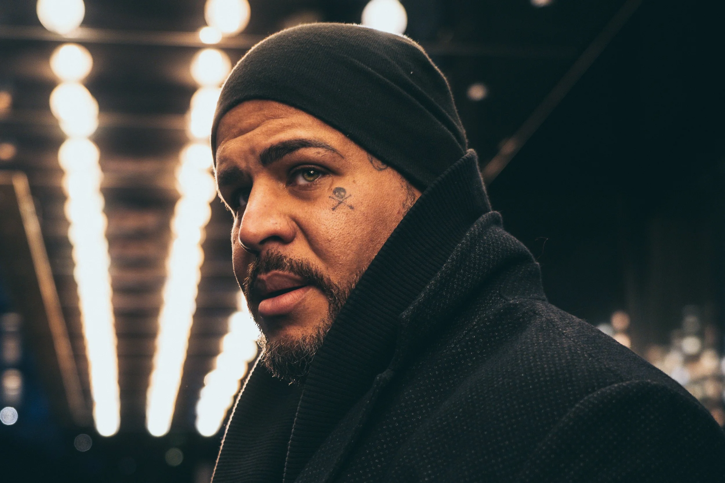 A man wearing a black beanie and black coat looking at the camera with a tattoo of a skull and crossbones on his cheek, in an indoor space with bright lights in the background.