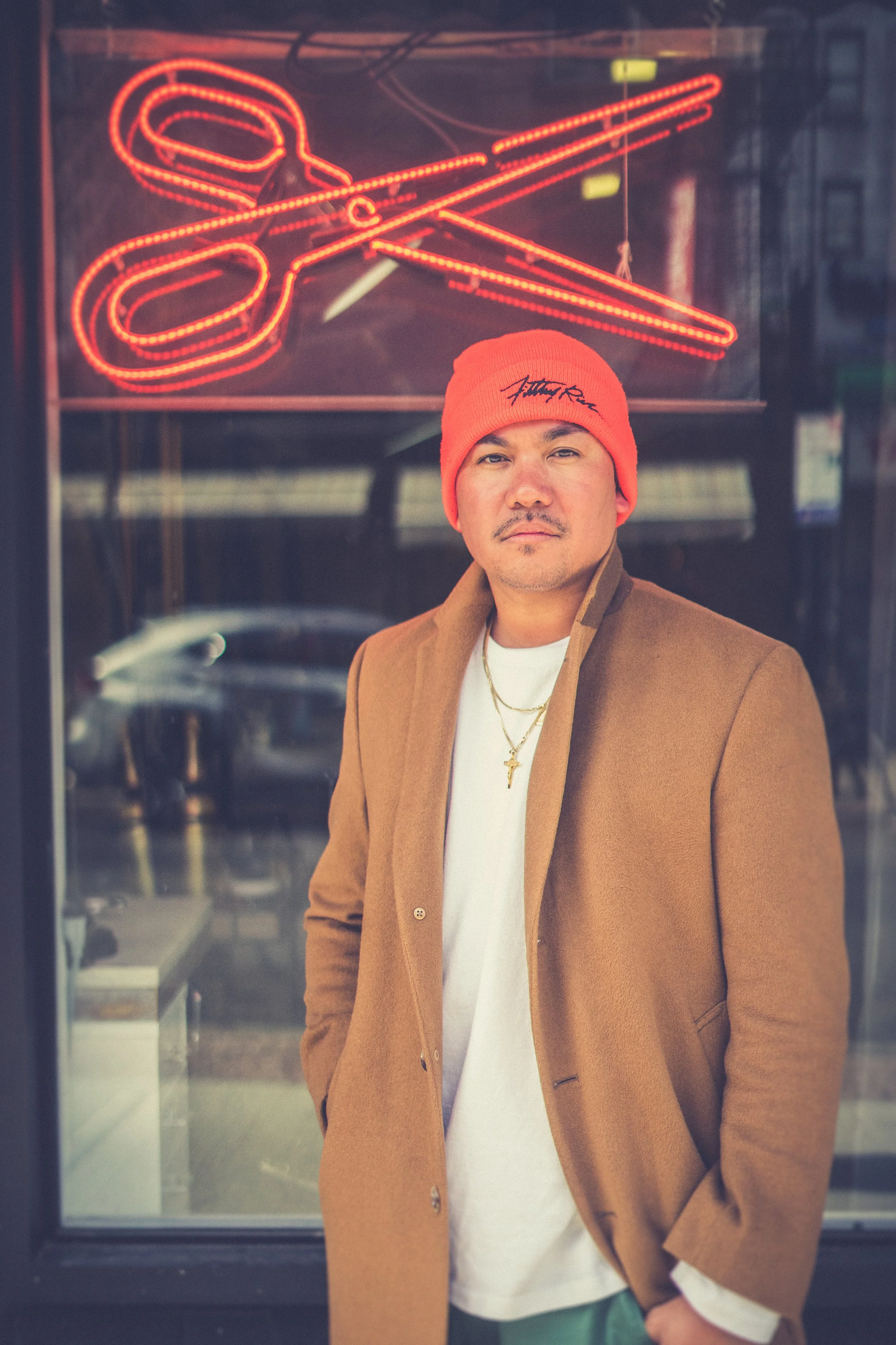A man wearing a red beanie, white shirt, gold necklaces, and a tan coat standing outside a store with a neon scissor sign in the background.