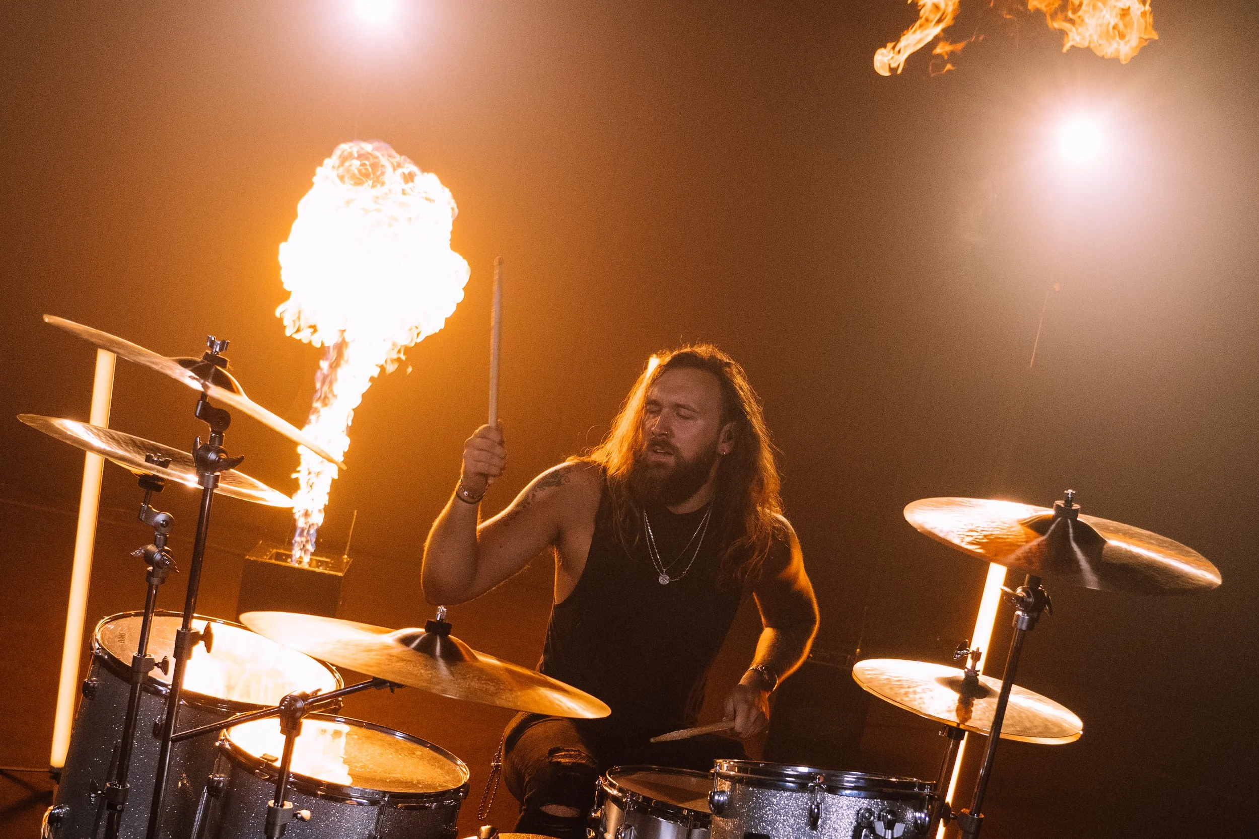 A drummer with long hair and a beard performing on stage, with flames and bright lights in the background.