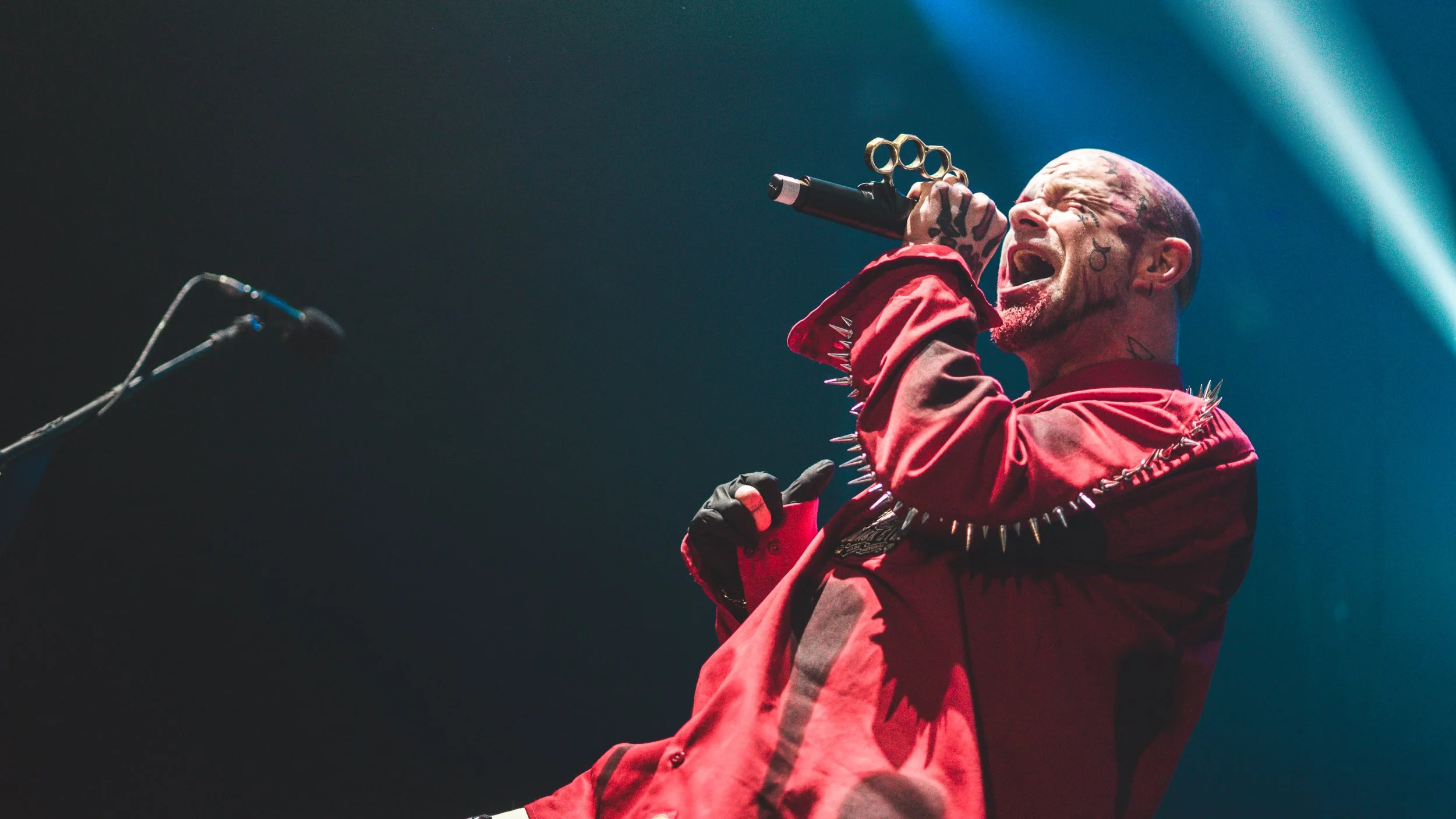 A male singer with tattoos on his face and head, wearing a red jacket with metal spikes on the shoulder, is passionately performing with a microphone in his hand on a dark stage with blue lights.