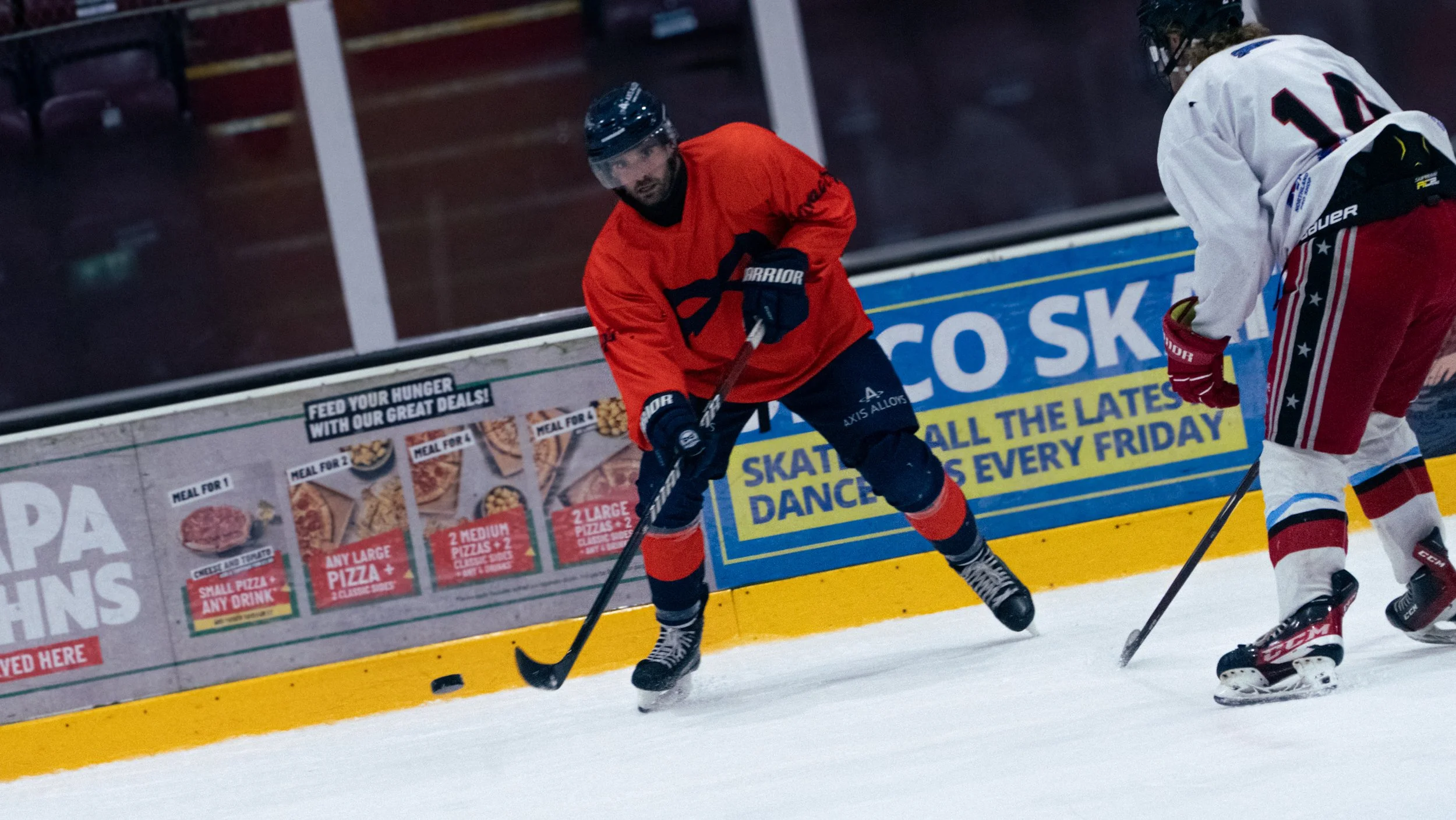 Two hockey players in action on the ice rink during a game. One player is wearing a red jersey, black pants, and a helmet, while the other is in a white jersey with red, black, and white socks. They are competing for the puck near the boards.