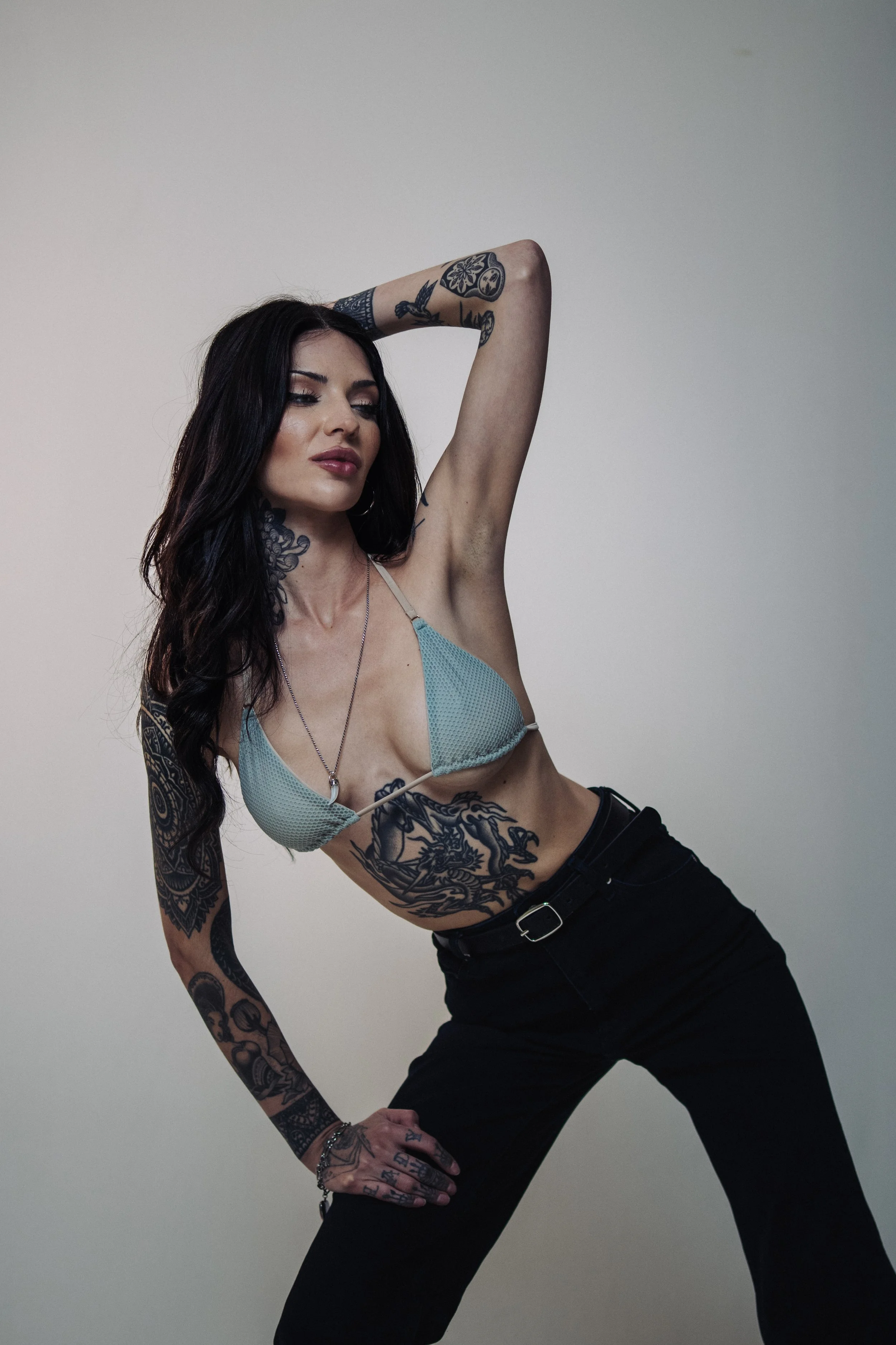 A woman with long dark hair, tattoos on arms, neck, and side, wearing a light blue bra and black pants, posing with one arm behind her head against a plain neutral background.