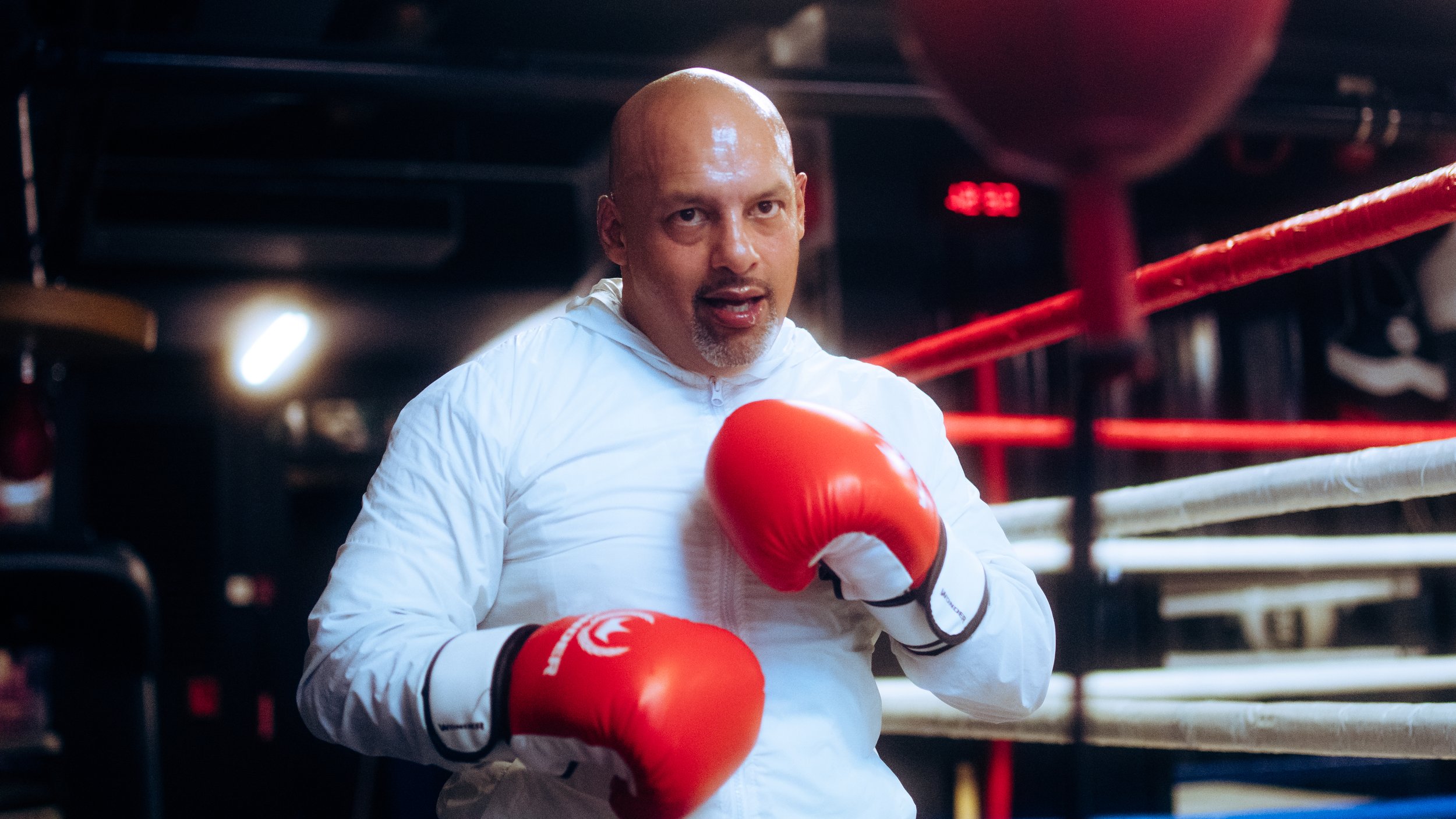 A man with a shaved head and goatee wearing red boxing gloves and a white jacket in a boxing ring.
