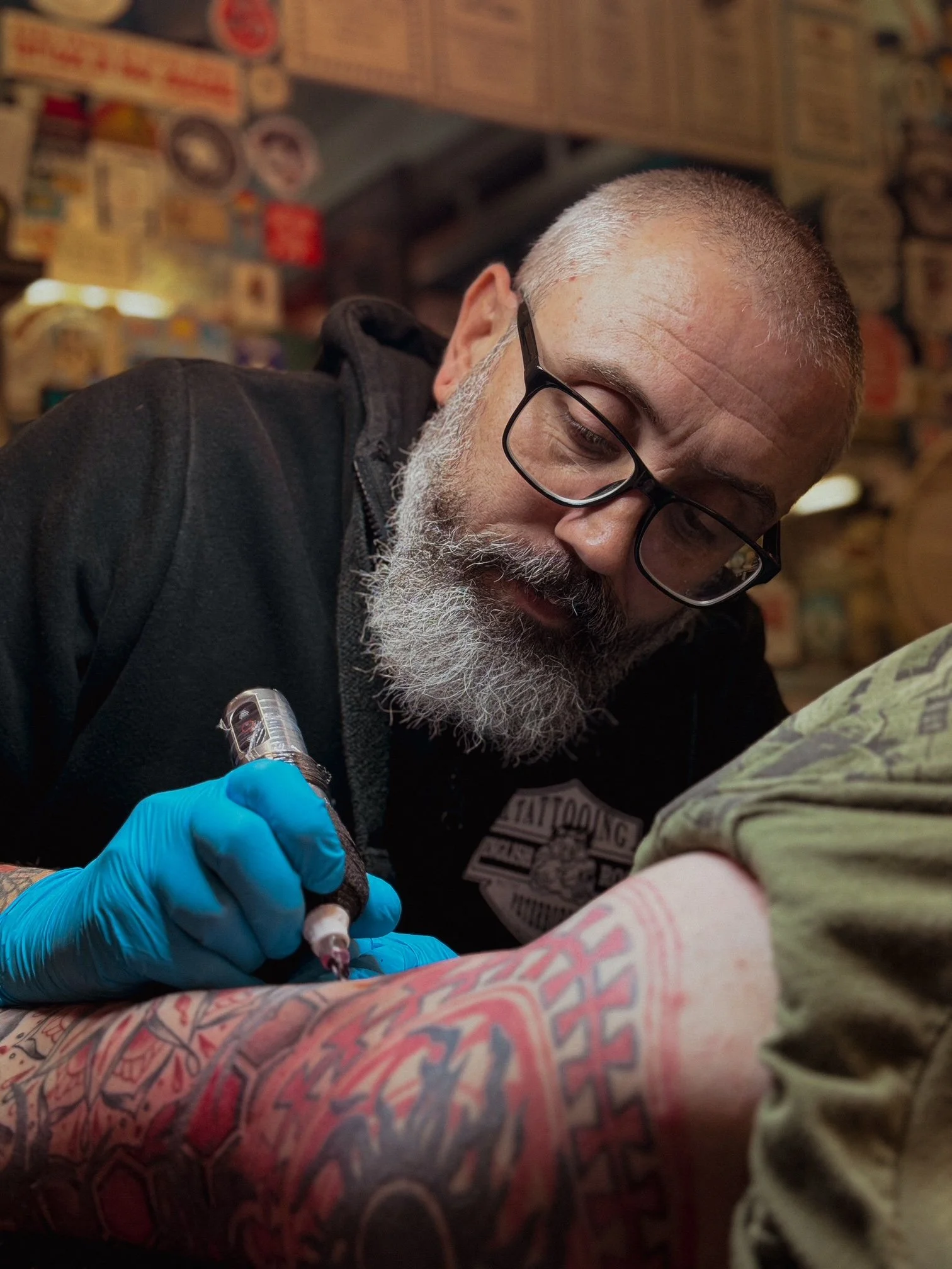 A man with glasses and a gray beard is tattooing a colorful tattoo on a person's arm in a tattoo parlor, with tattoos and posters on the wall in the background.