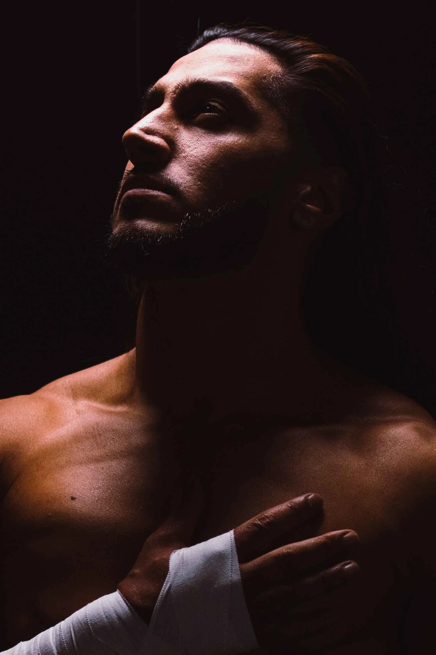 A muscular man with long hair and a beard poses with his hand on his chest, in a dark background with dramatic lighting highlighting his face and upper body.