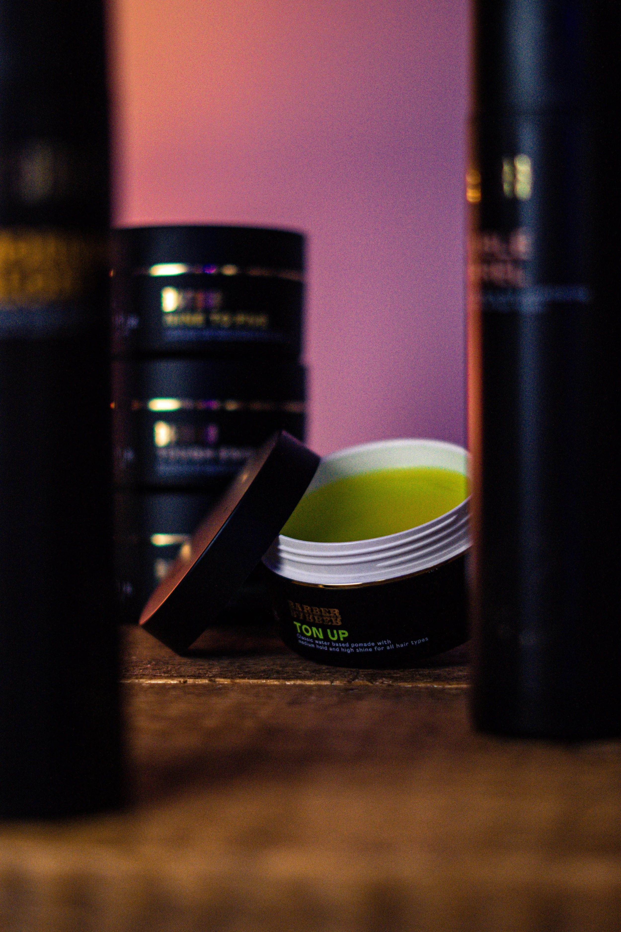 Open black container of hair pomade on a wooden surface, surrounded by other hair product containers.