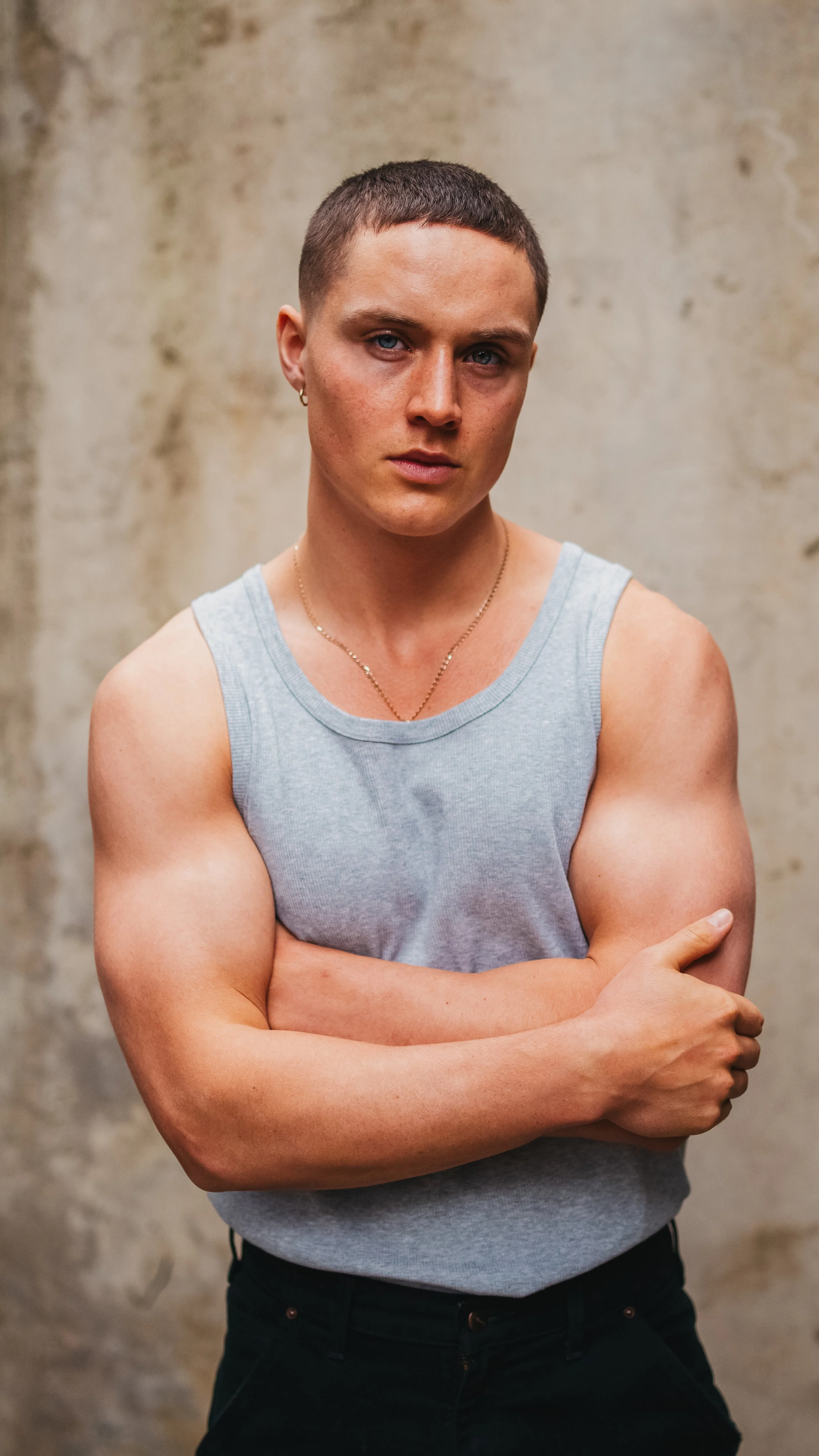 A woman with short hair wearing a gray tank top, black pants, and jewelry, standing against a neutral background with her arms crossed, looking at the camera.