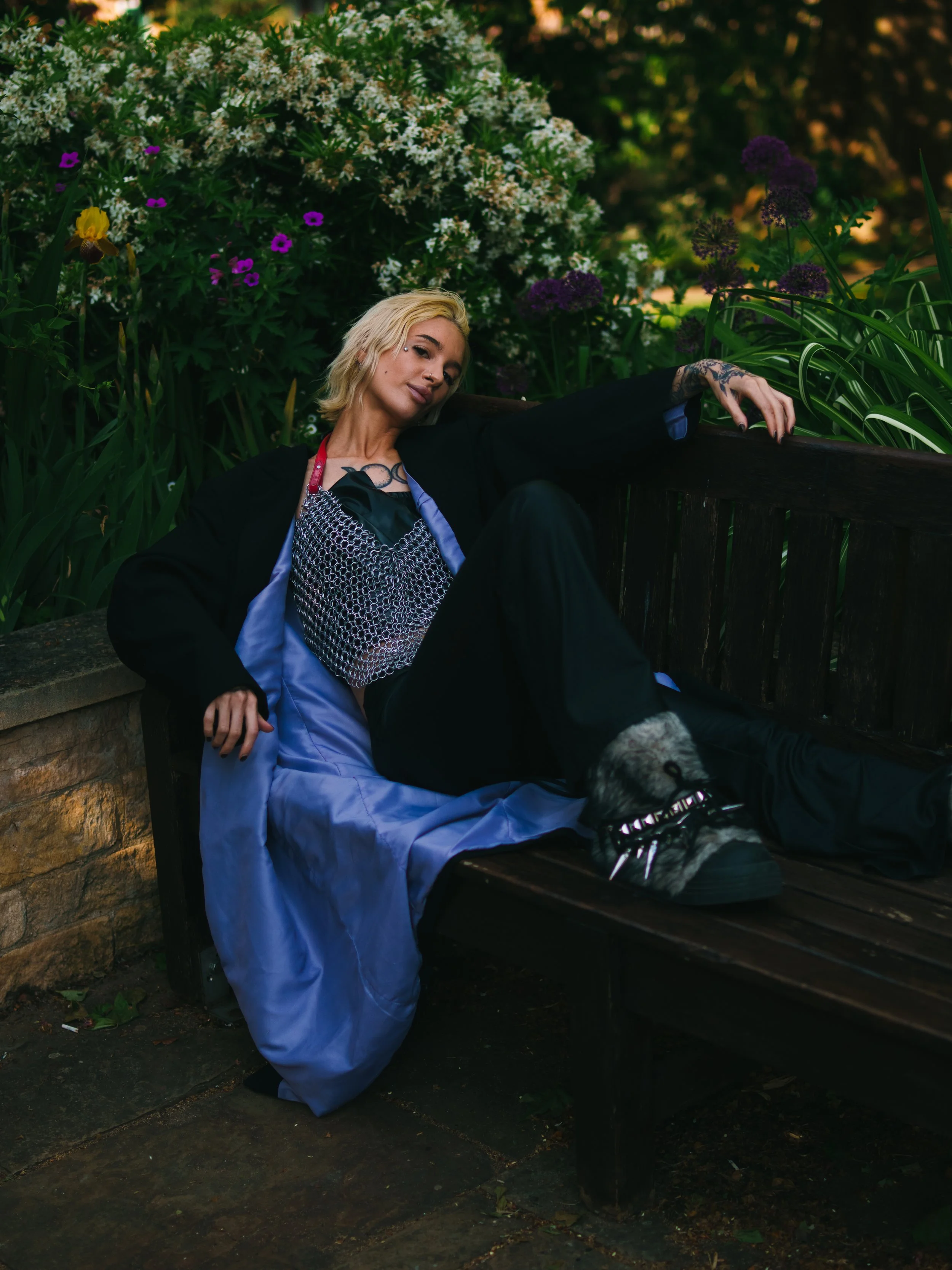 A woman with blonde hair relaxing on a park bench surrounded by lush green plants and flowers, wearing a black and purple outfit, with tattoos on her arm, and wearing a shoe with a dog face design.