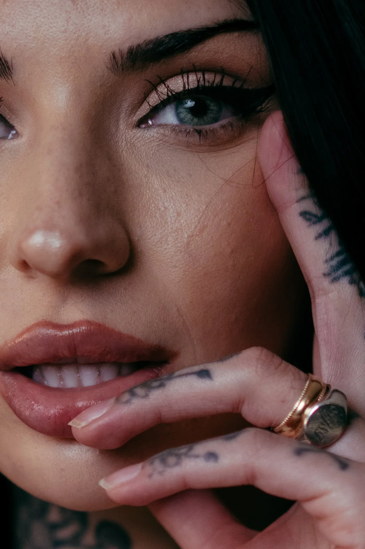 Close-up of a woman's face, showing one eye, lips, and tattooed fingers. She has dark hair, makeup, and wears rings on her fingers.
