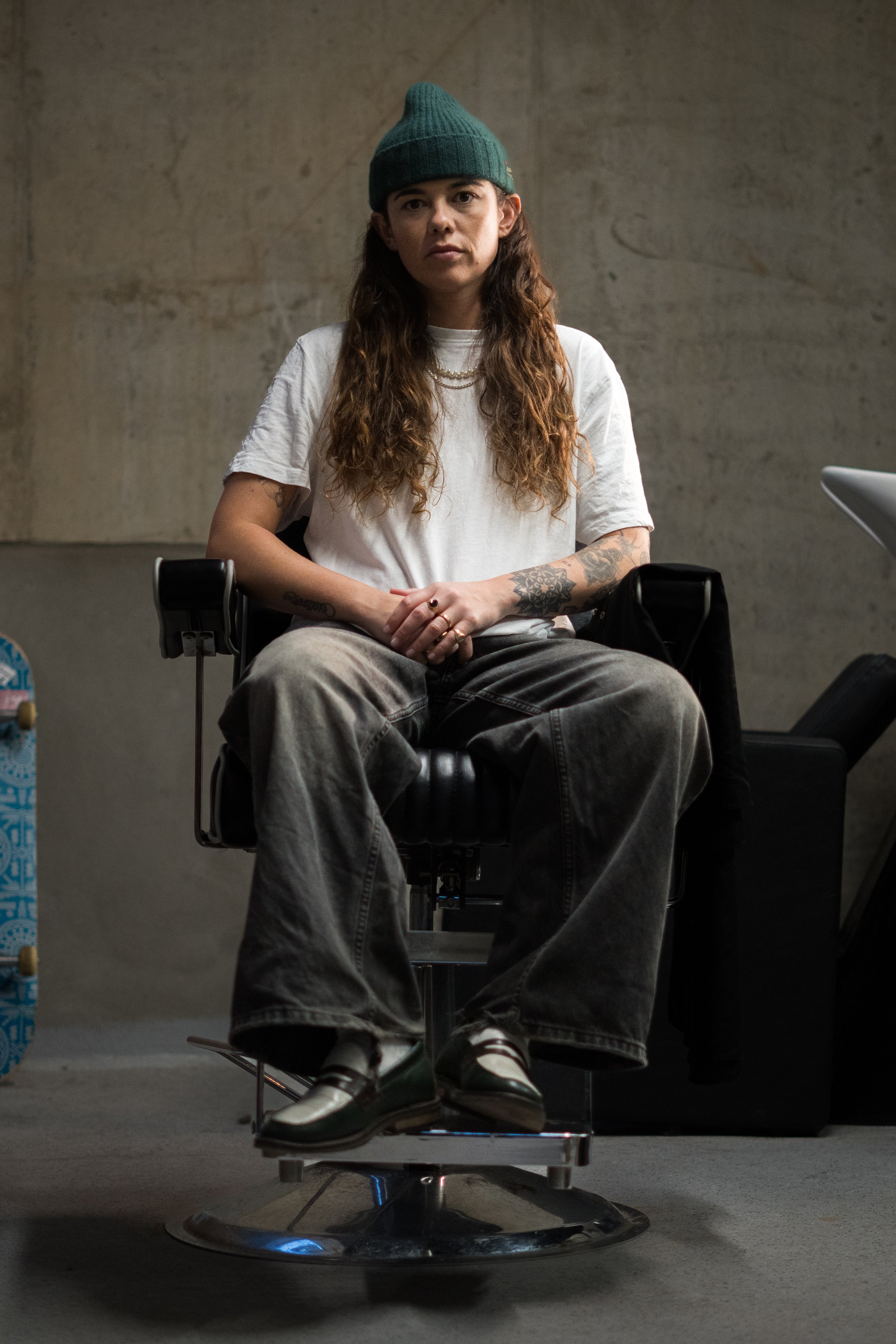 A young woman with long brown hair, tattoos, wearing a teal beanie, white t-shirt, and gray baggy jeans, sitting in a black barber chair with a gray concrete wall background.