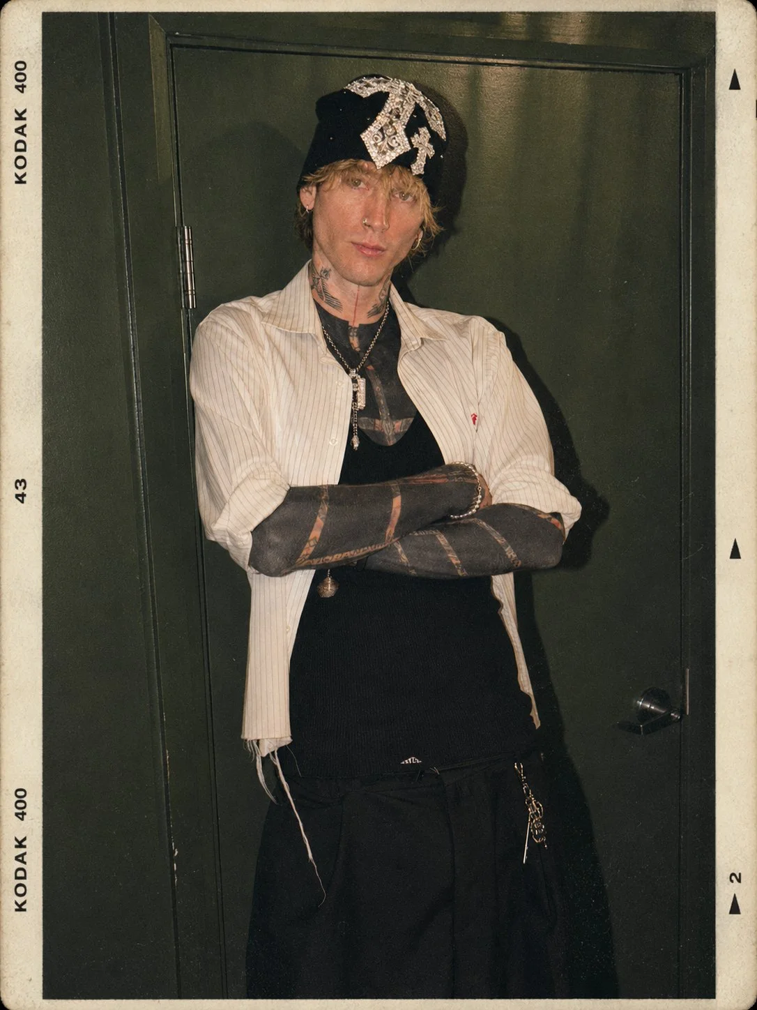 Young person with tattoos and jewelry, wearing a black beanie with embellishments, striped shirt over a black shirt, and black pants, standing with arms crossed against a dark green door.