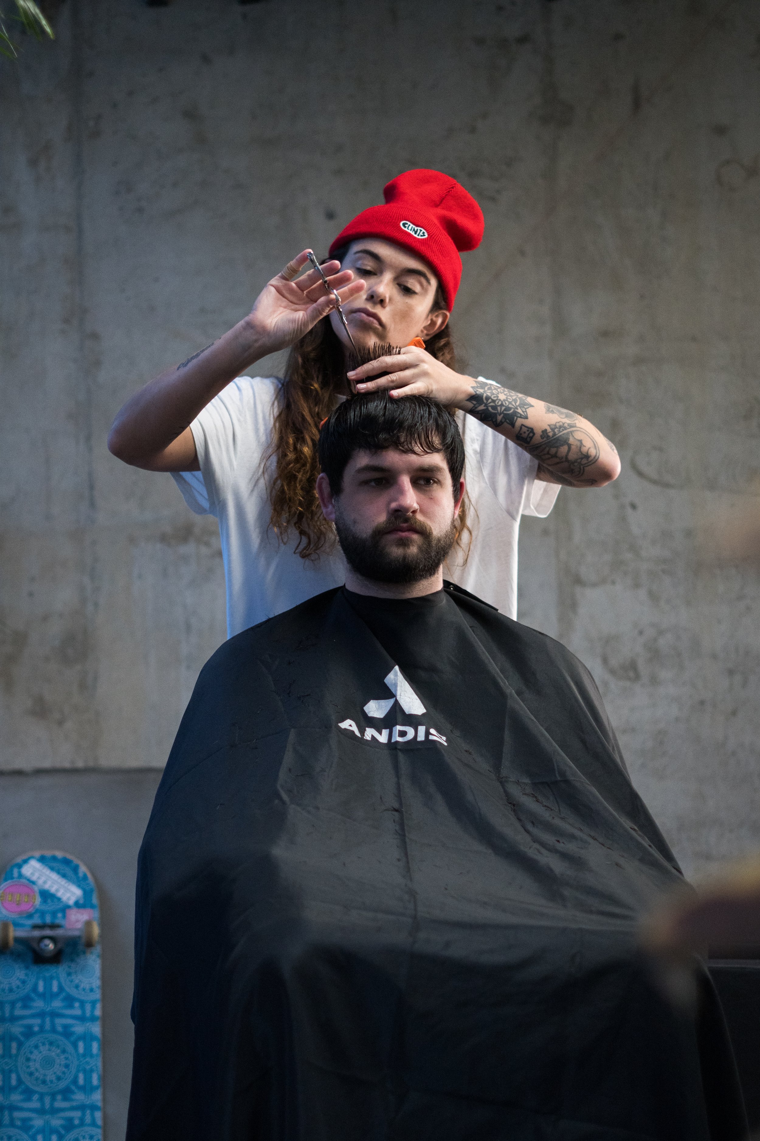 A woman with tattoos and a red beanie haircutting a man's hair in a barbershop.