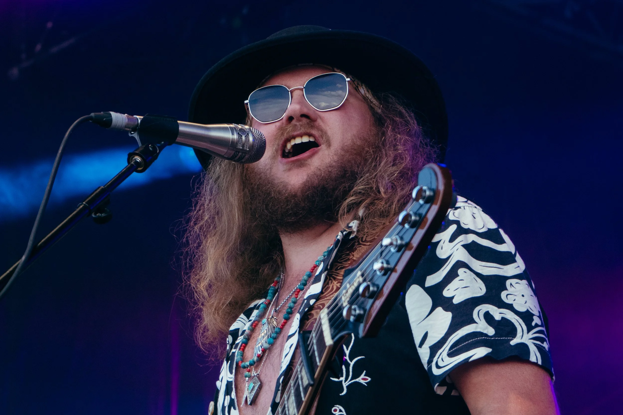 Male musician with long hair and beard singing into microphone, wearing sunglasses, black hat, and patterned shirt, playing guitar during a live performance.