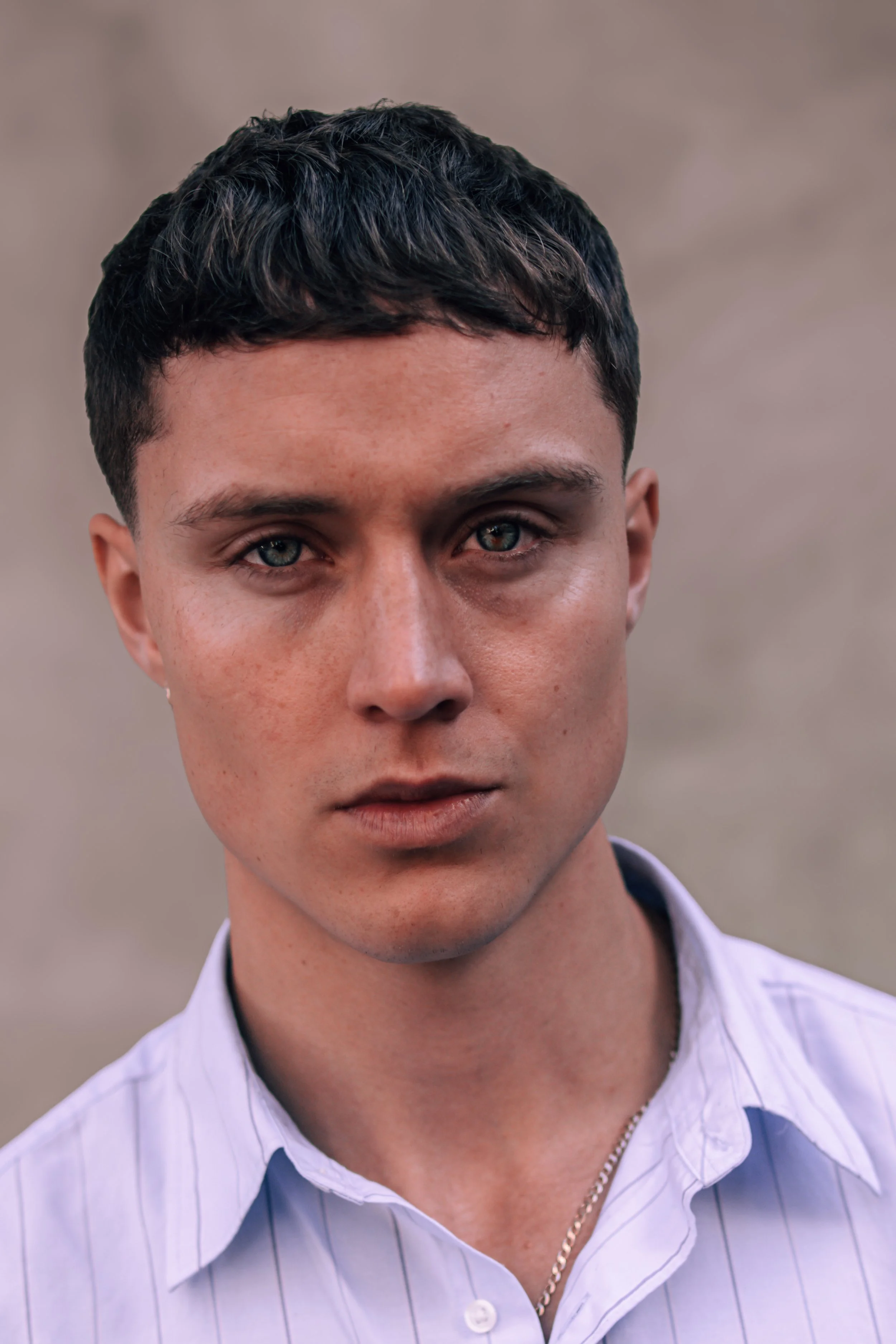 A young man with short dark hair and blue eyes, wearing a light purple striped shirt, has a serious expression and is looking directly at the camera. The background is neutral and blurred.