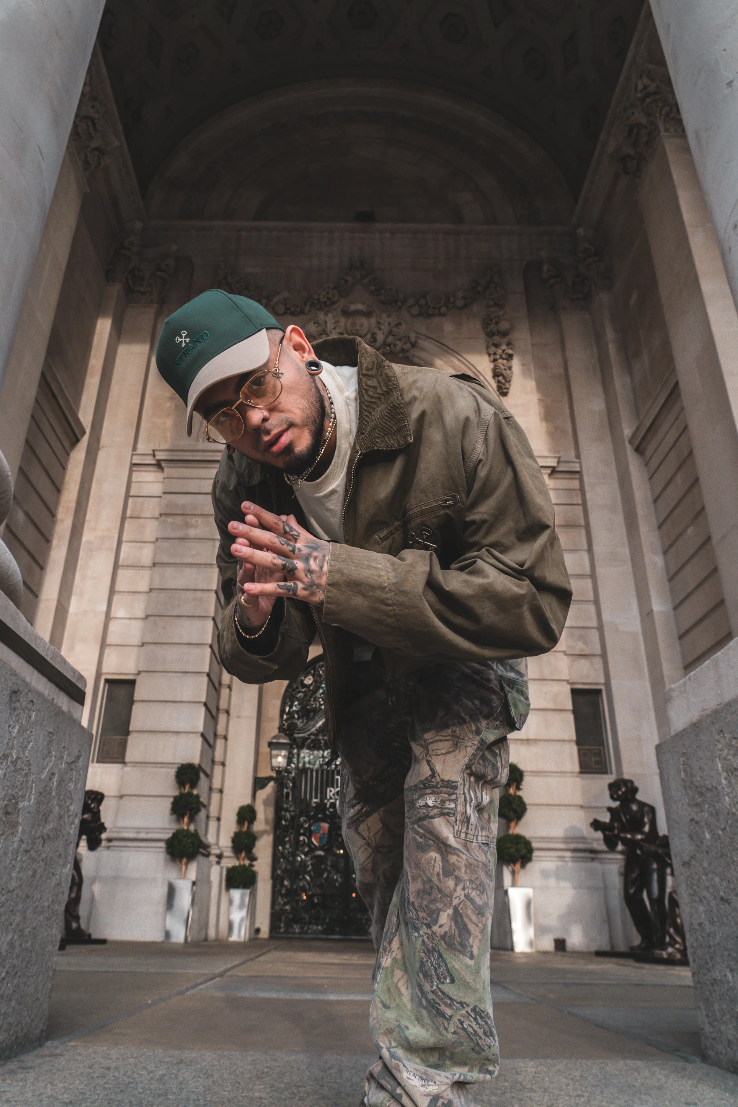 A man with tattoos, sunglasses, and piercings poses in front of an ornate building with statues and potted plants, wearing a green cap, sunglasses, camouflage pants, and a brown jacket.