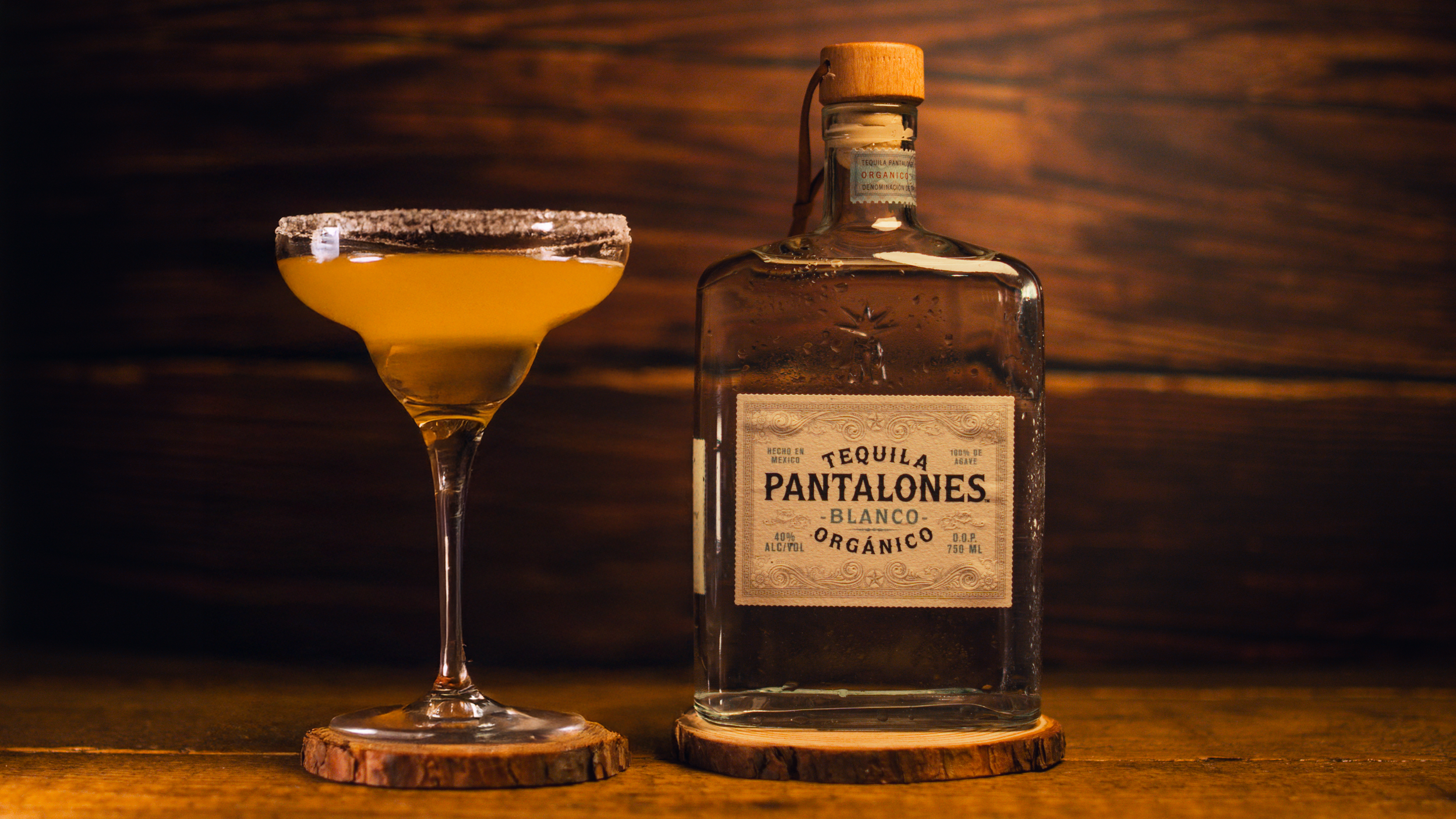 A bottle of tequila Pantalones Blanco Orgánico with a wooden cap next to a cocktail glass filled with yellow drink, both placed on wooden coasters on a wooden surface with a dark wooden background.