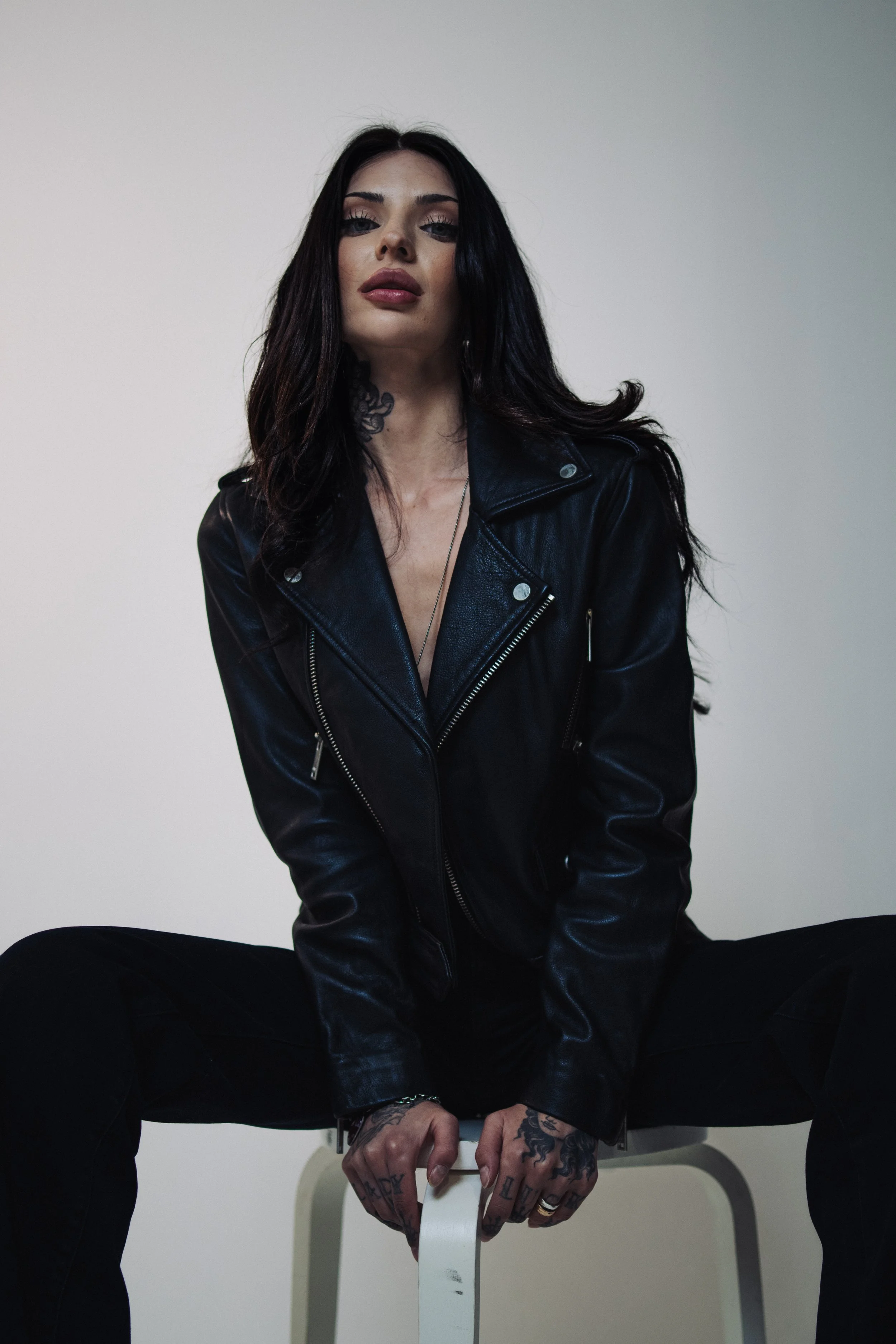 A young woman with long dark hair, tattoos on her hands and neck, sitting on a small white stool against a plain white wall, wearing a black leather jacket and black pants.