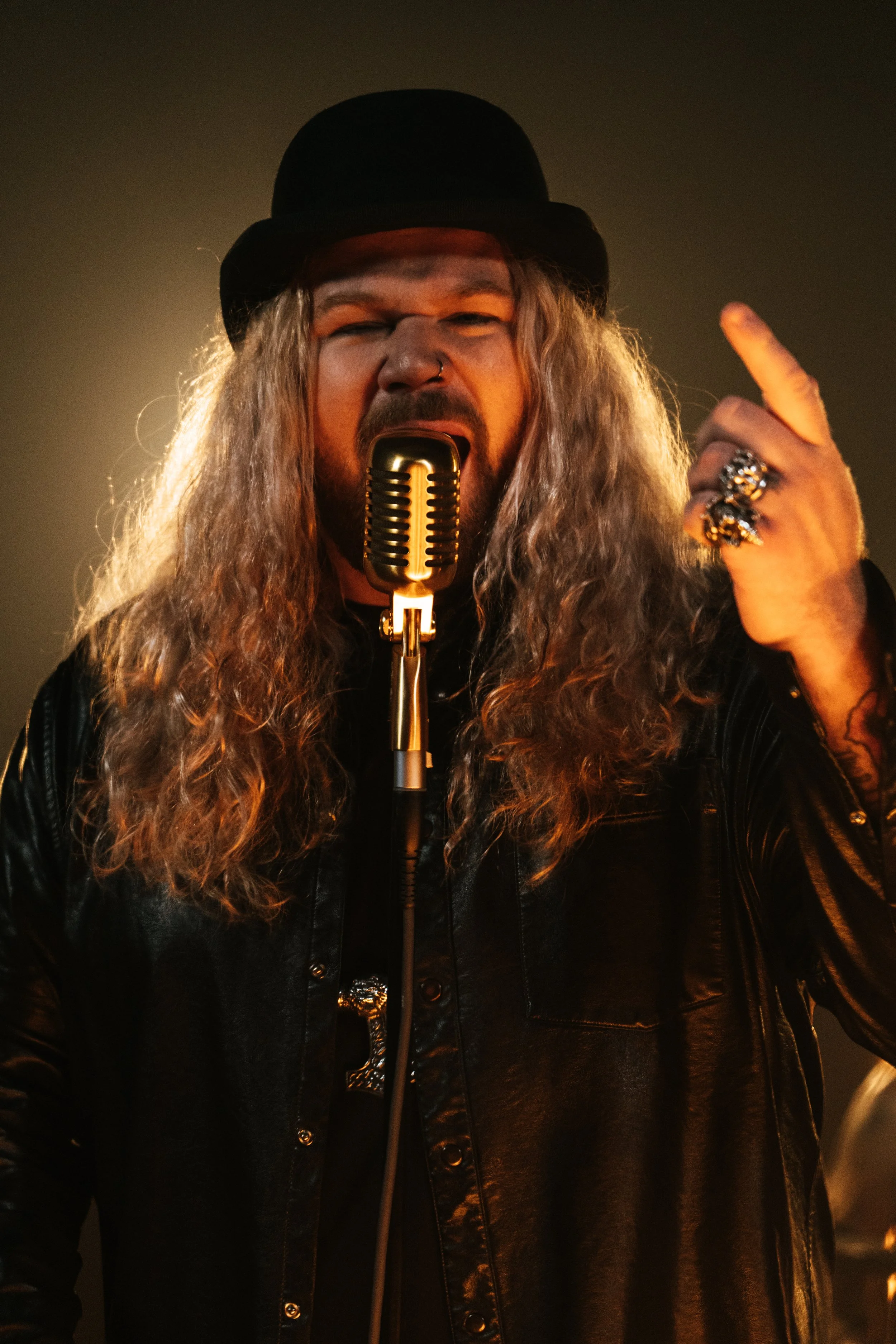 A male singer with long curly hair wearing a black shirt, rings, and a black hat, singing into a vintage microphone with one finger raised.