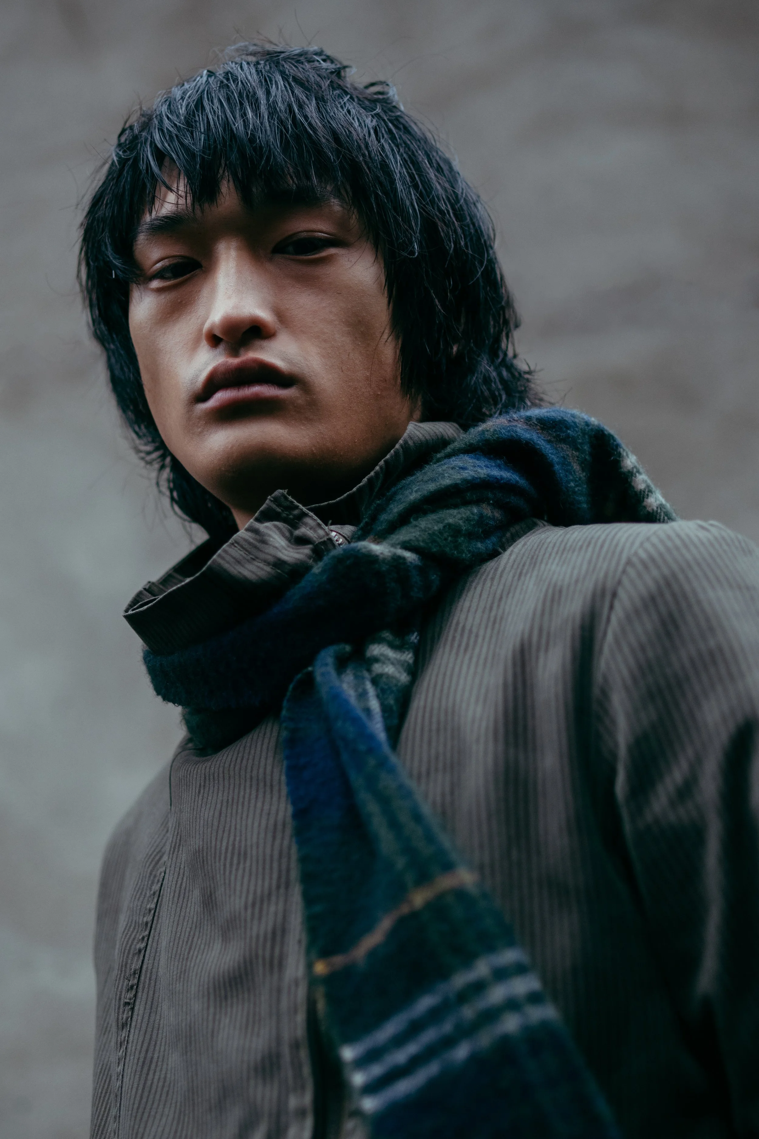 A young man with medium-length black hair and a serious expression, wearing a brown jacket and a dark blue scarf, standing outdoors against a cloudy sky.