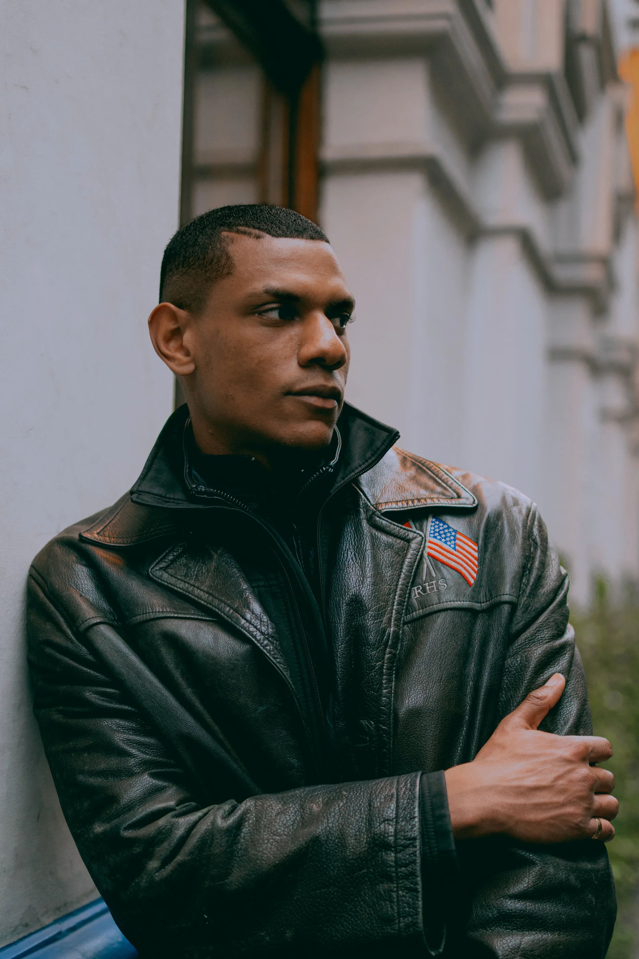 A young man with short hair wearing a black leather jacket with an American flag patch, standing against a wall with a city street in the background.