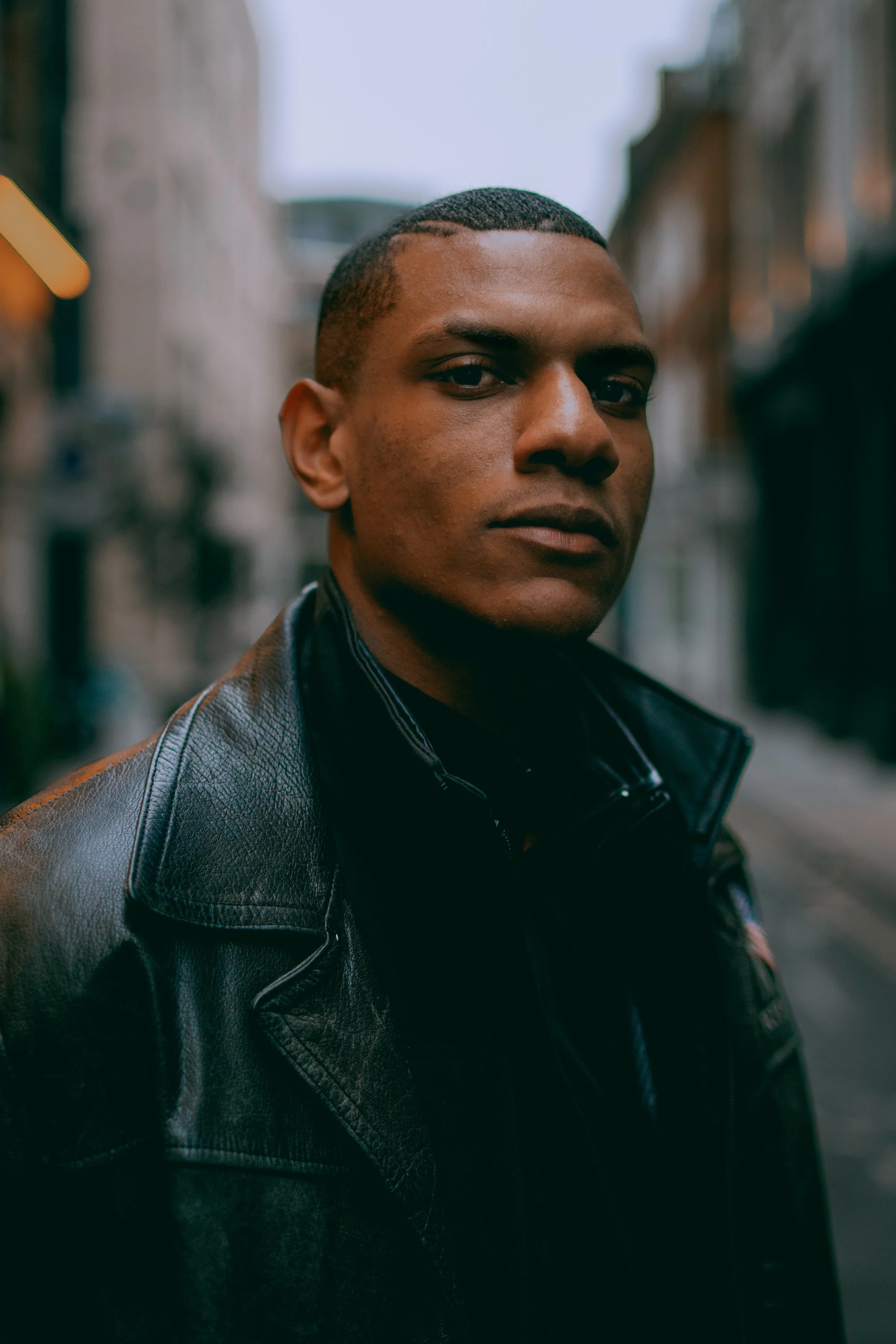A young man with short hair wearing a black leather jacket outdoors in an urban setting.