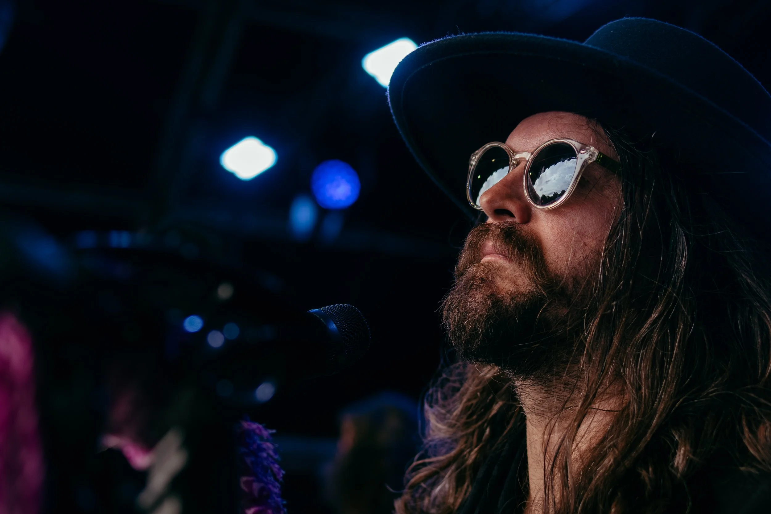 A man with long hair, a beard, wearing sunglasses and a wide-brimmed hat, is performing in a dark setting with stage lights reflecting off his sunglasses.