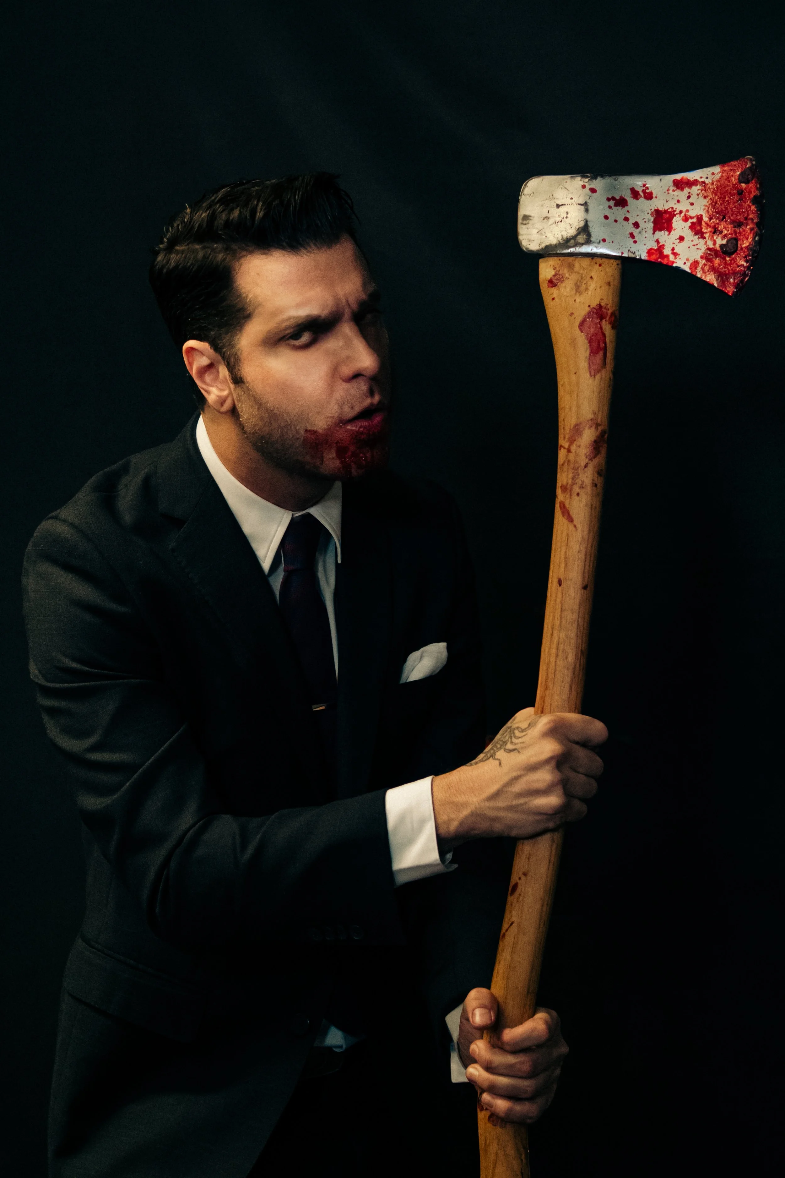 A man in a black suit with a tie and white shirt holding a bloodstained axe, with blood on his face and lips, against a dark background.