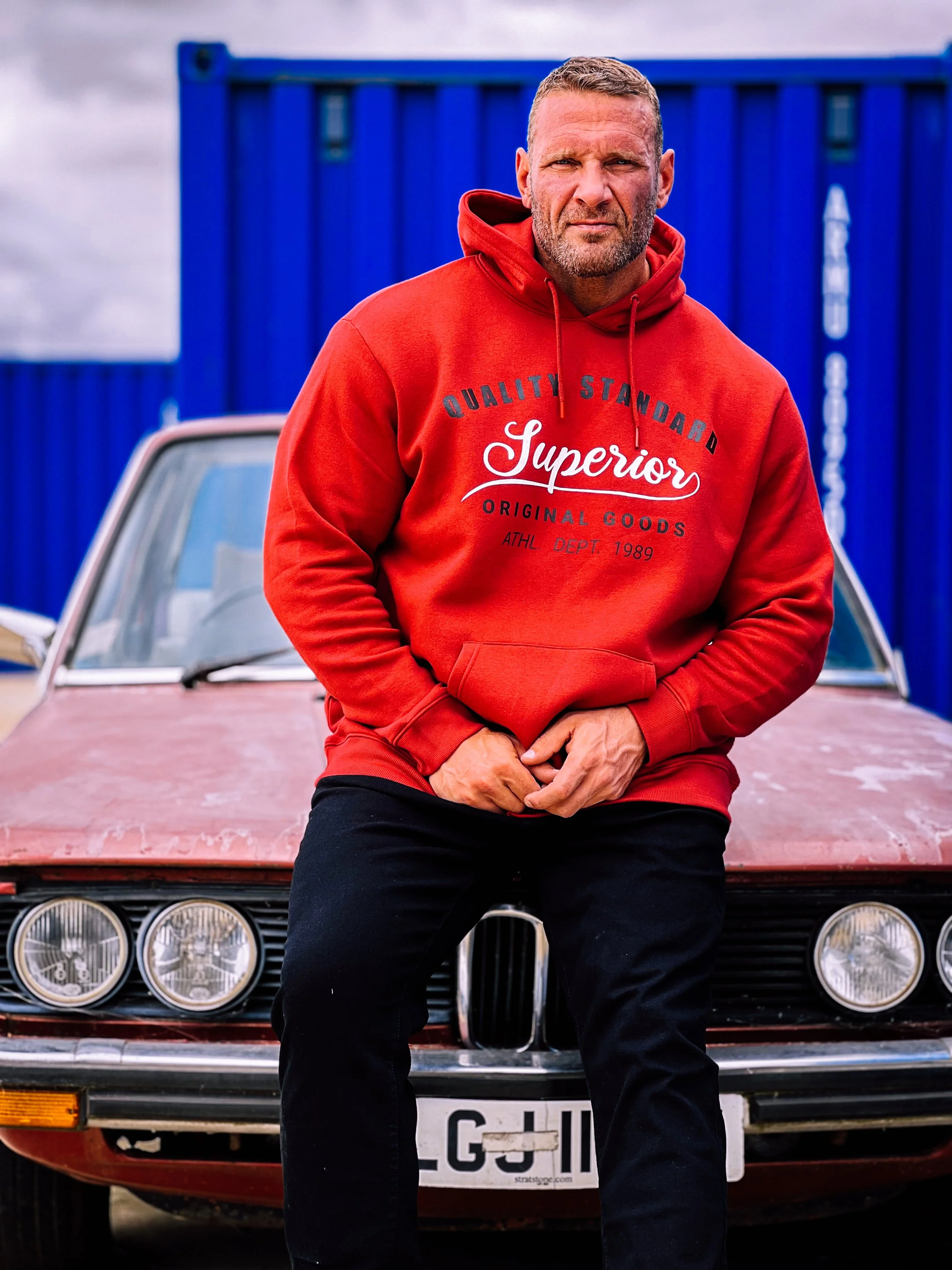 A man in a red hoodie with text, standing in front of an old red car with a blue fence in the background.