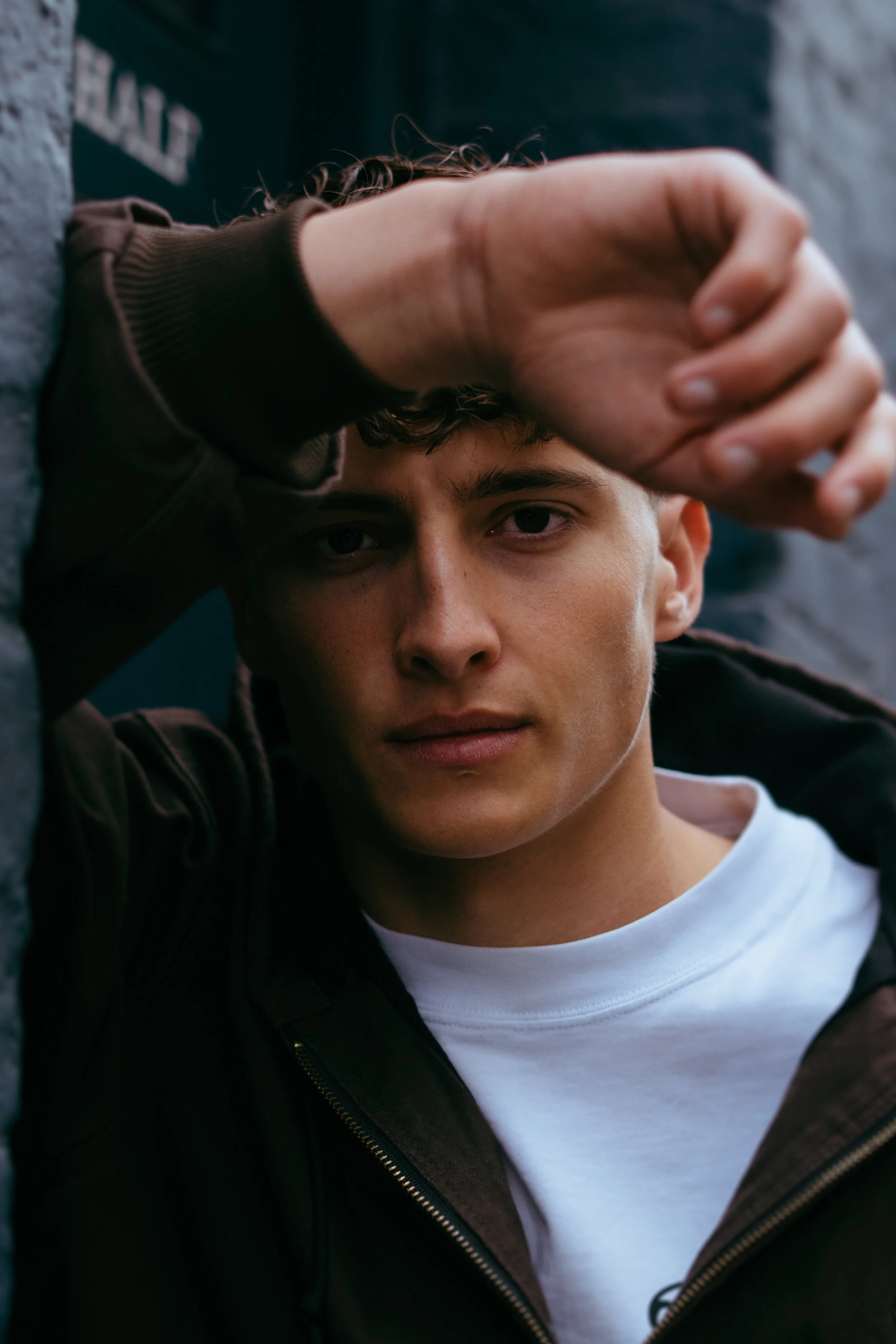 Young man lying down, resting his head on a stone surface with his arm above his forehead, wearing a dark jacket over a white t-shirt