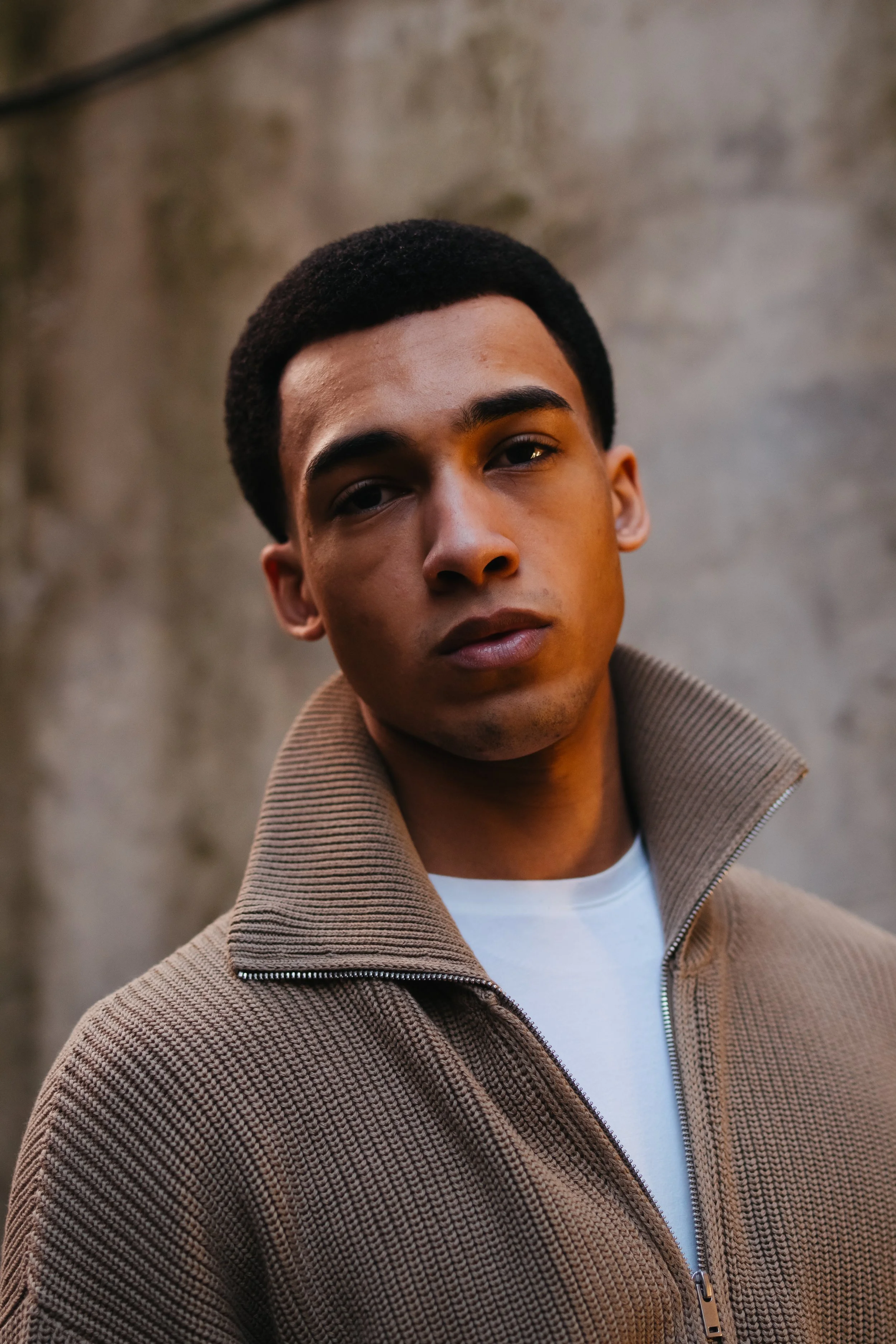 Portrait of a young man with short black hair wearing a tan zip-up jacket over a white shirt, standing against a blurred stone wall background.