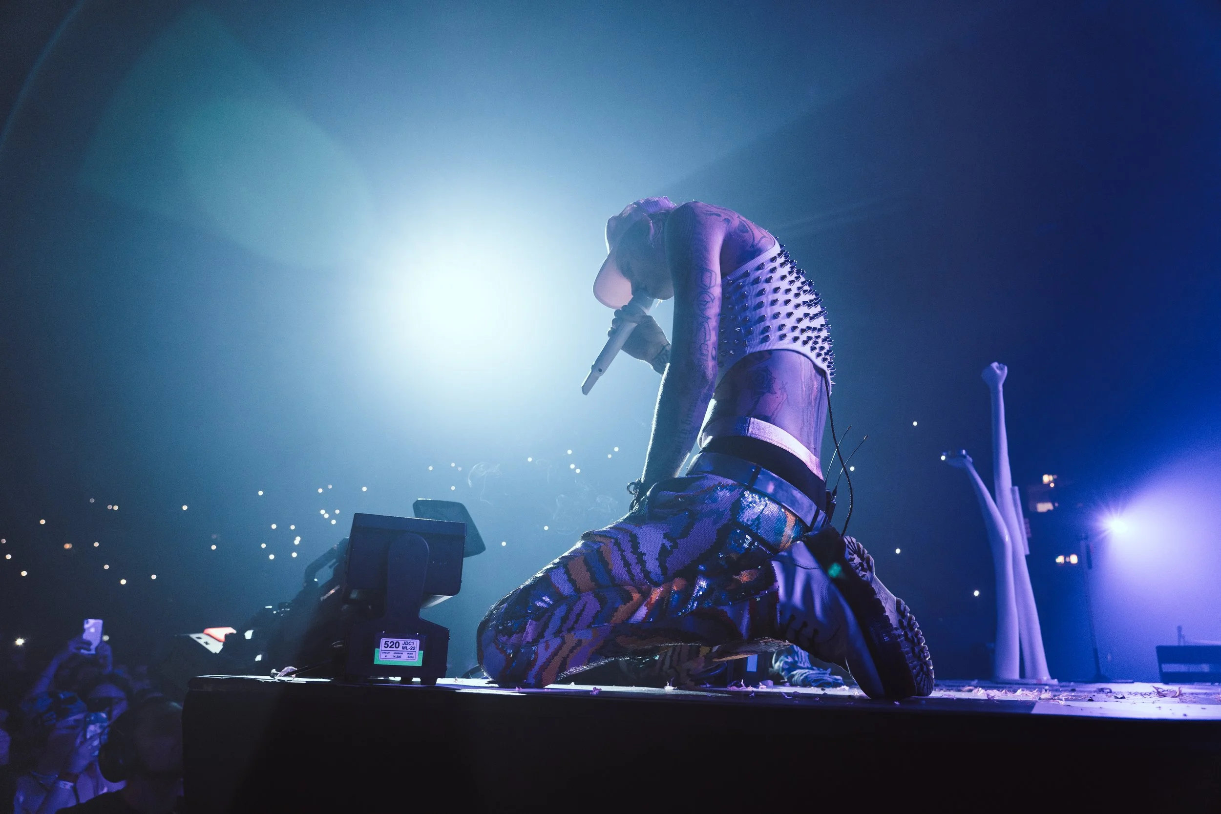 A performer is kneeling on stage, holding a microphone and bowing their head with eyes closed. They are wearing a studded sleeveless top, colorful patterned pants, and a hat. Bright stage lights cast a glow behind them, and audience members are visib