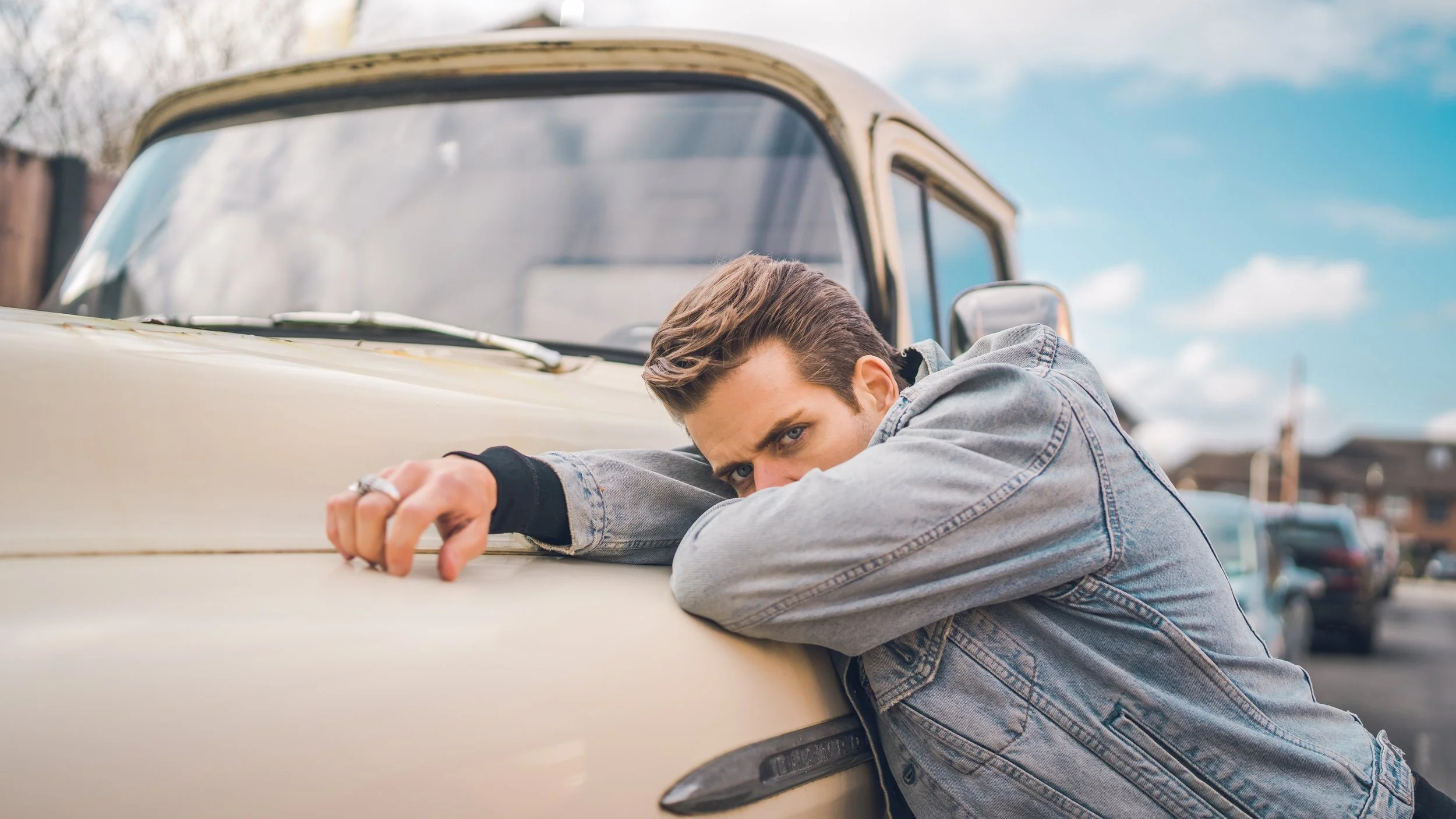 A young man leaning on a vintage cream-colored car, resting his head and arms on the hood, with a serious expression and piercing eyes, outdoors with a cloudy blue sky and cars in the background.