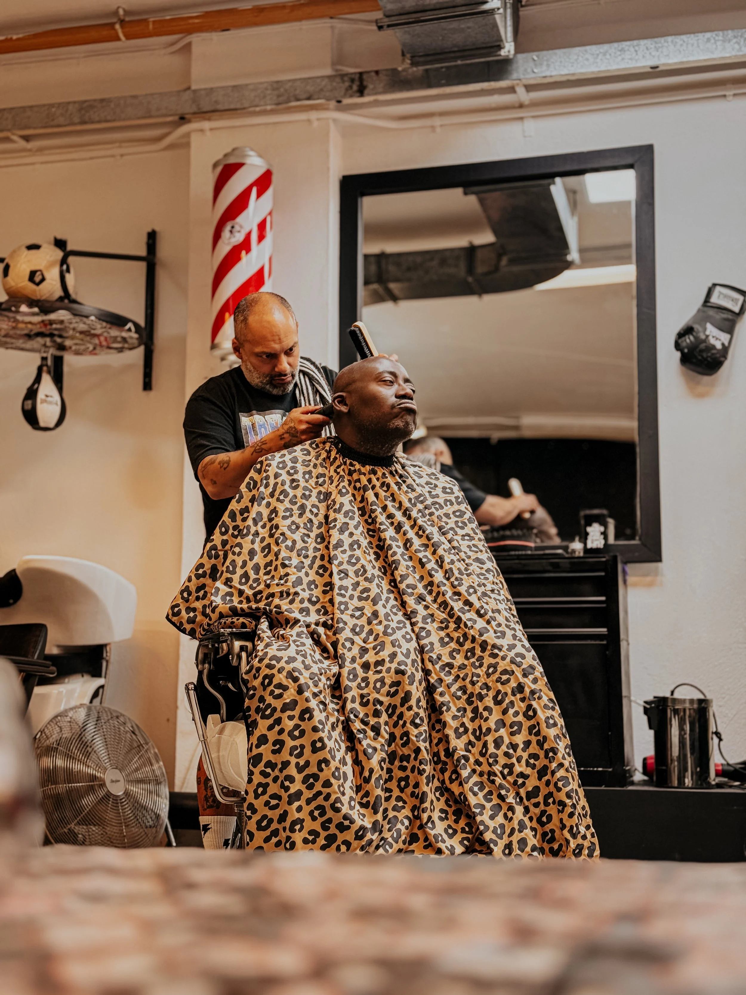A man getting a haircut at a barbershop, wearing a leopard print cape. The barber is standing behind him, cutting his hair. The barbershop has various equipment, a mirror, and decorative elements.