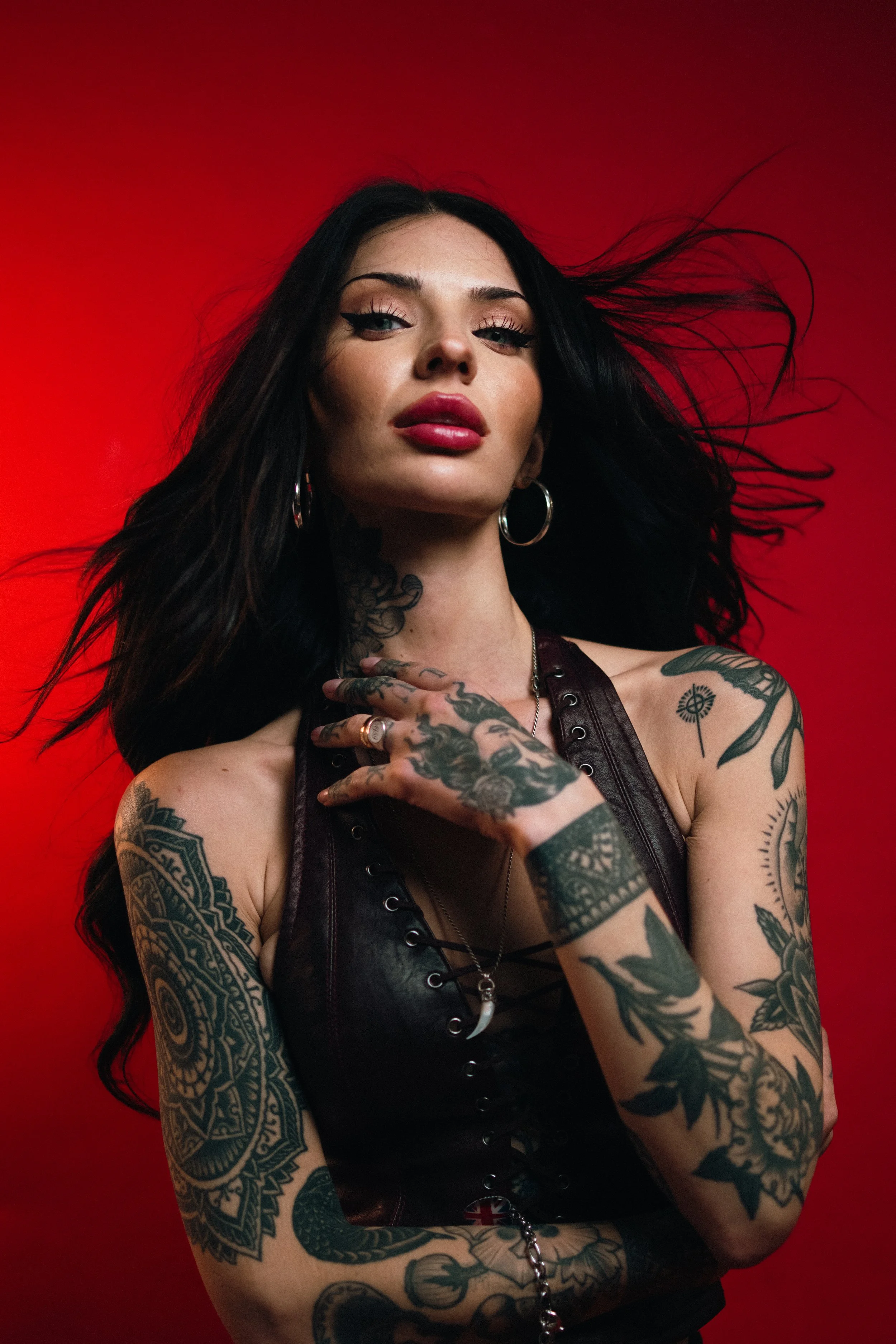 A woman with long black hair, tattoos on her arms and neck, wearing hoop earrings and a black leather top, poses against a red background.