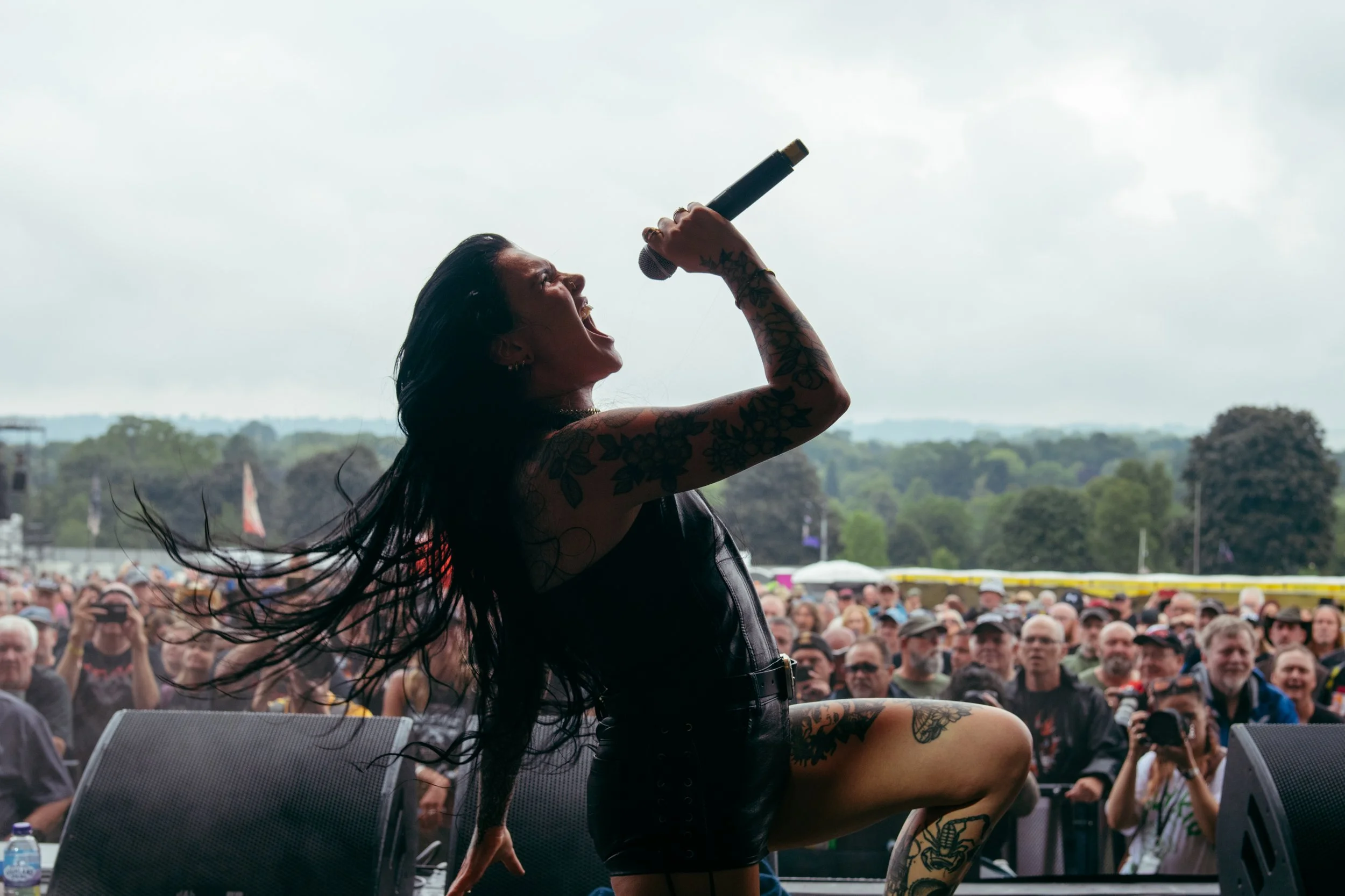 A female singer performs passionately on stage with a microphone in her hand, outdoors during a cloudy day, with a large crowd watching.