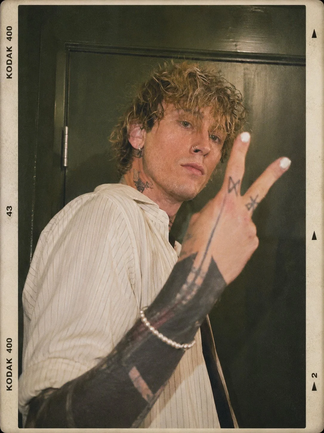 A young man with curly blond hair, tattoos, and piercings, wearing a cream-colored striped shirt and black gloves with tattoos, making a peace sign with his fingers.