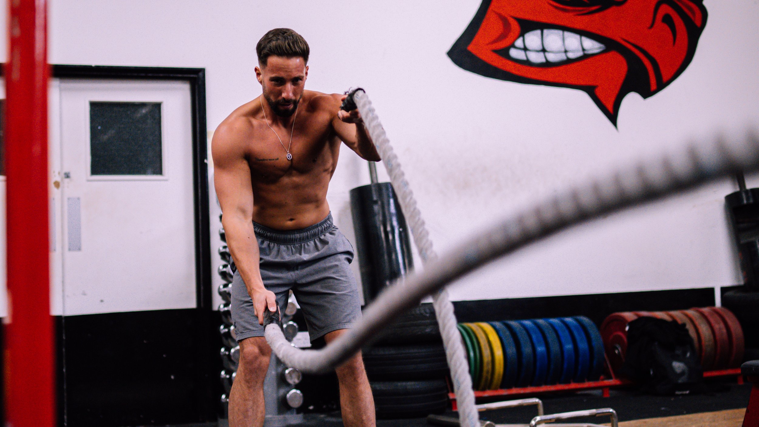 A shirtless man with gray shorts and a necklace exercises with battle ropes in a gym with colorful weight plates and a red and black logo on the wall.