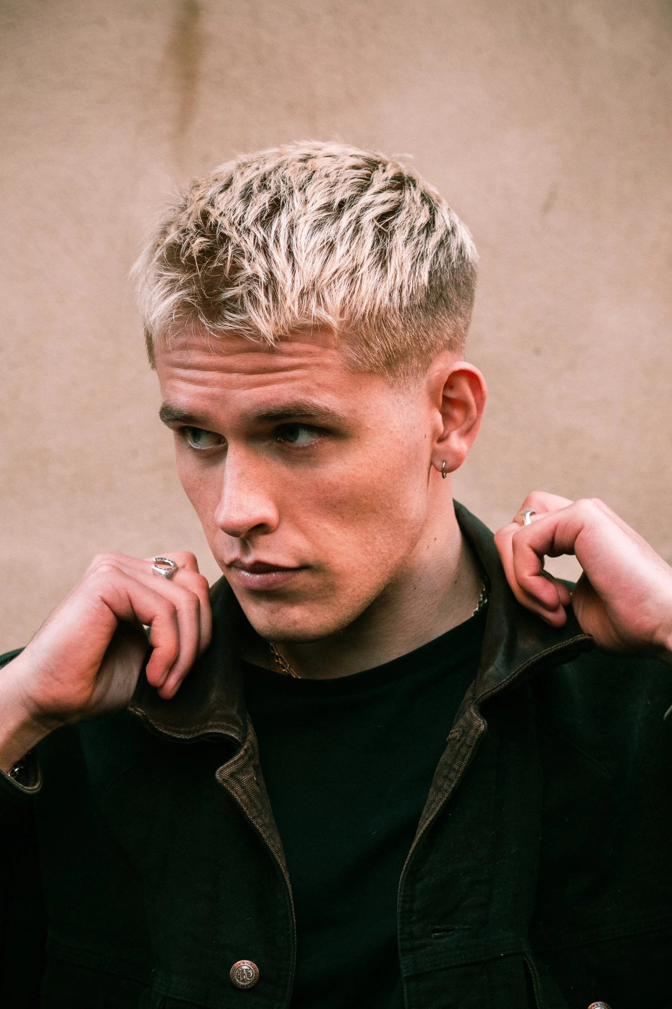 A young man with platinum blonde hair and a fade haircut, wearing earrings, a black shirt, and a dark jacket, adjusting his jacket collar.