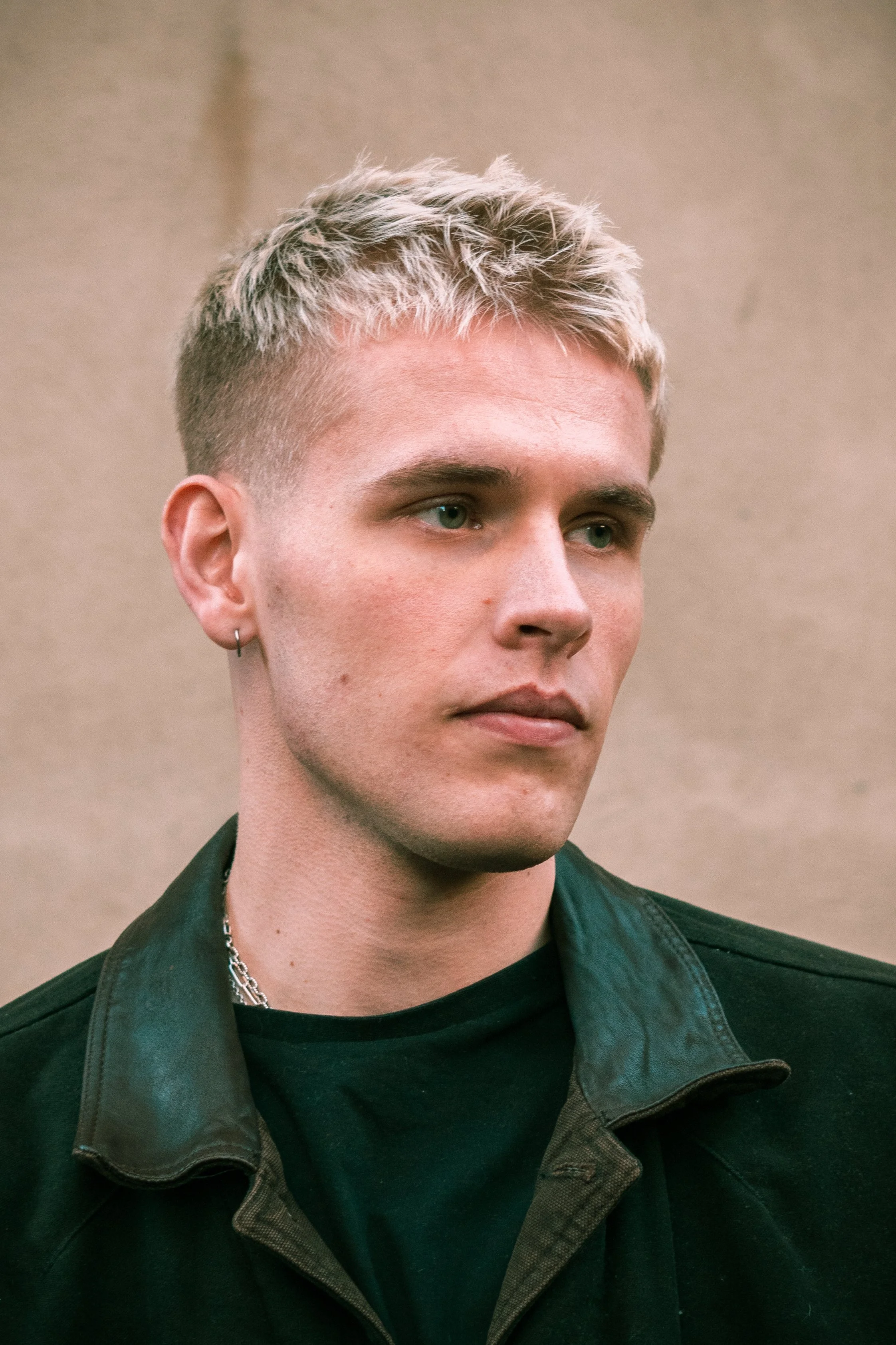 A young man with blonde hair, blue eyes, earrings, wearing a jacket, and a necklace, standing against a beige background.