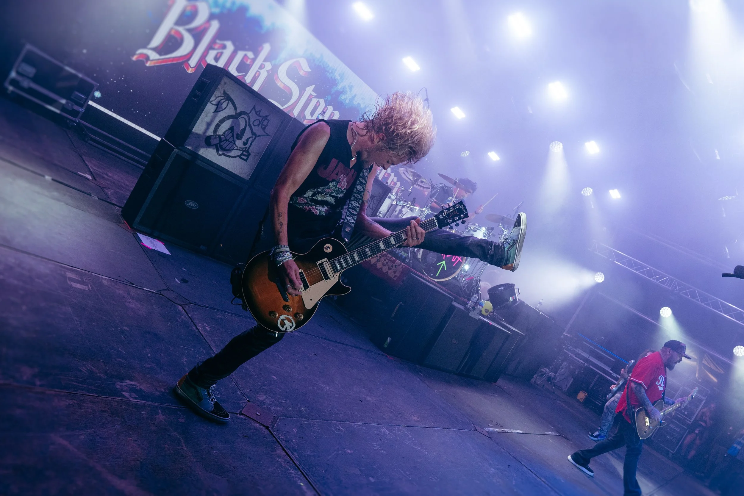 Musicians performing on stage with guitars, one musician is jumping with one leg up, surrounded by bright stage lights, a large Backstopp sign in the background.
