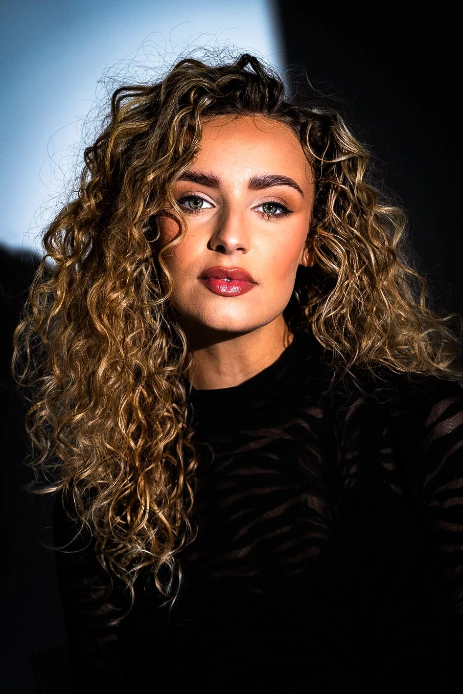 Close-up portrait of a woman with curly hair, wearing a black top, looking directly at the camera with a neutral expression against a dark background.