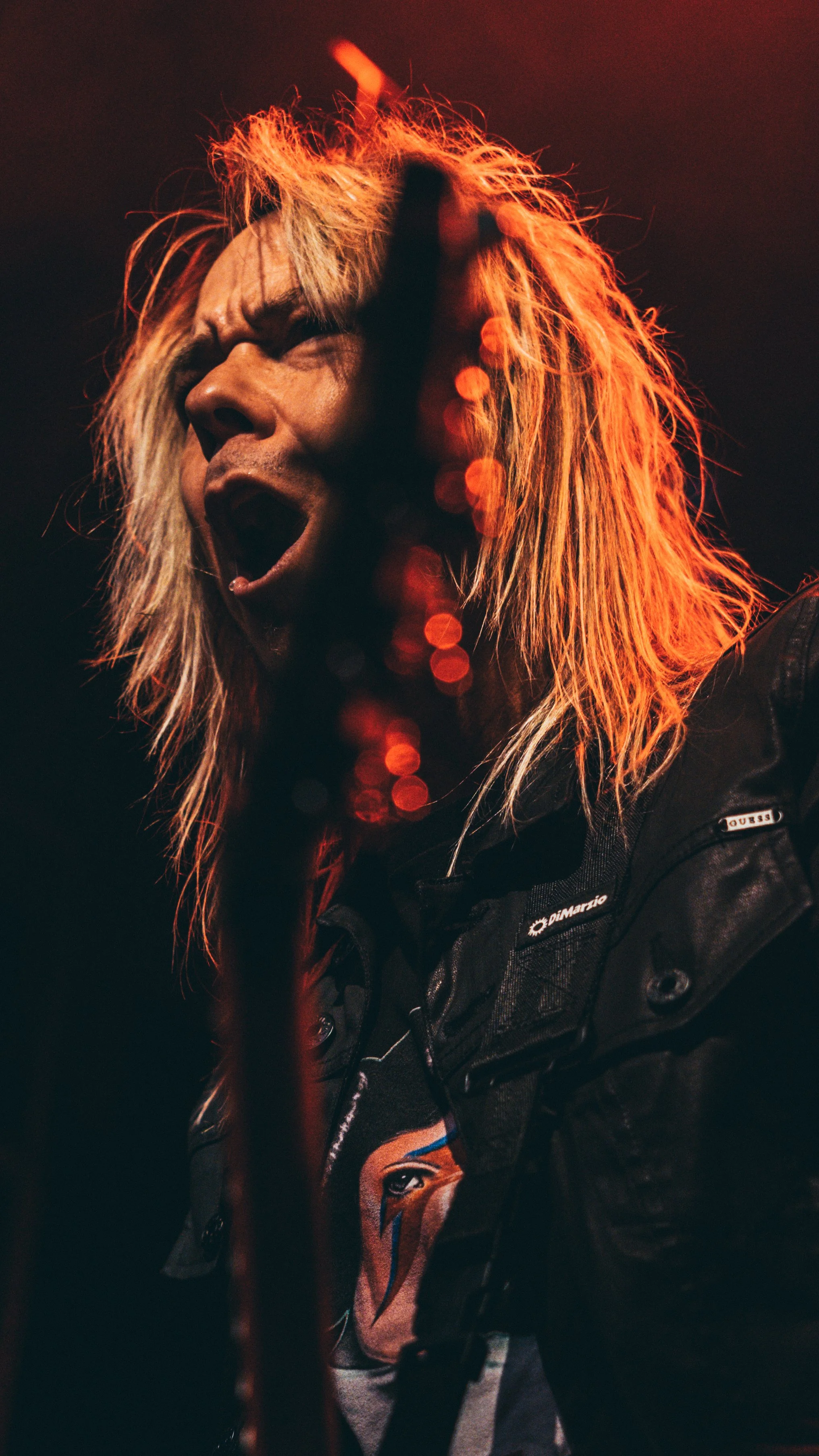 A singer with long, blond hair passionately performing on stage, with red and orange lighting creating a dramatic effect.