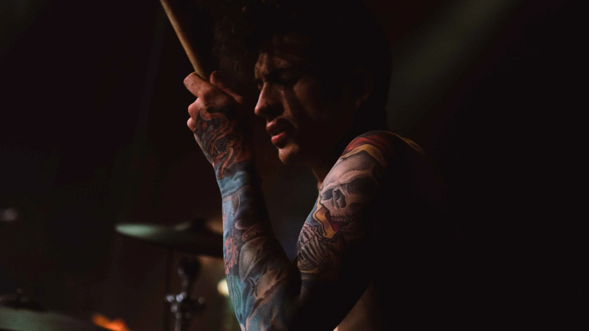 A tattooed man with dark hair, holding a cigar, in a dark setting.