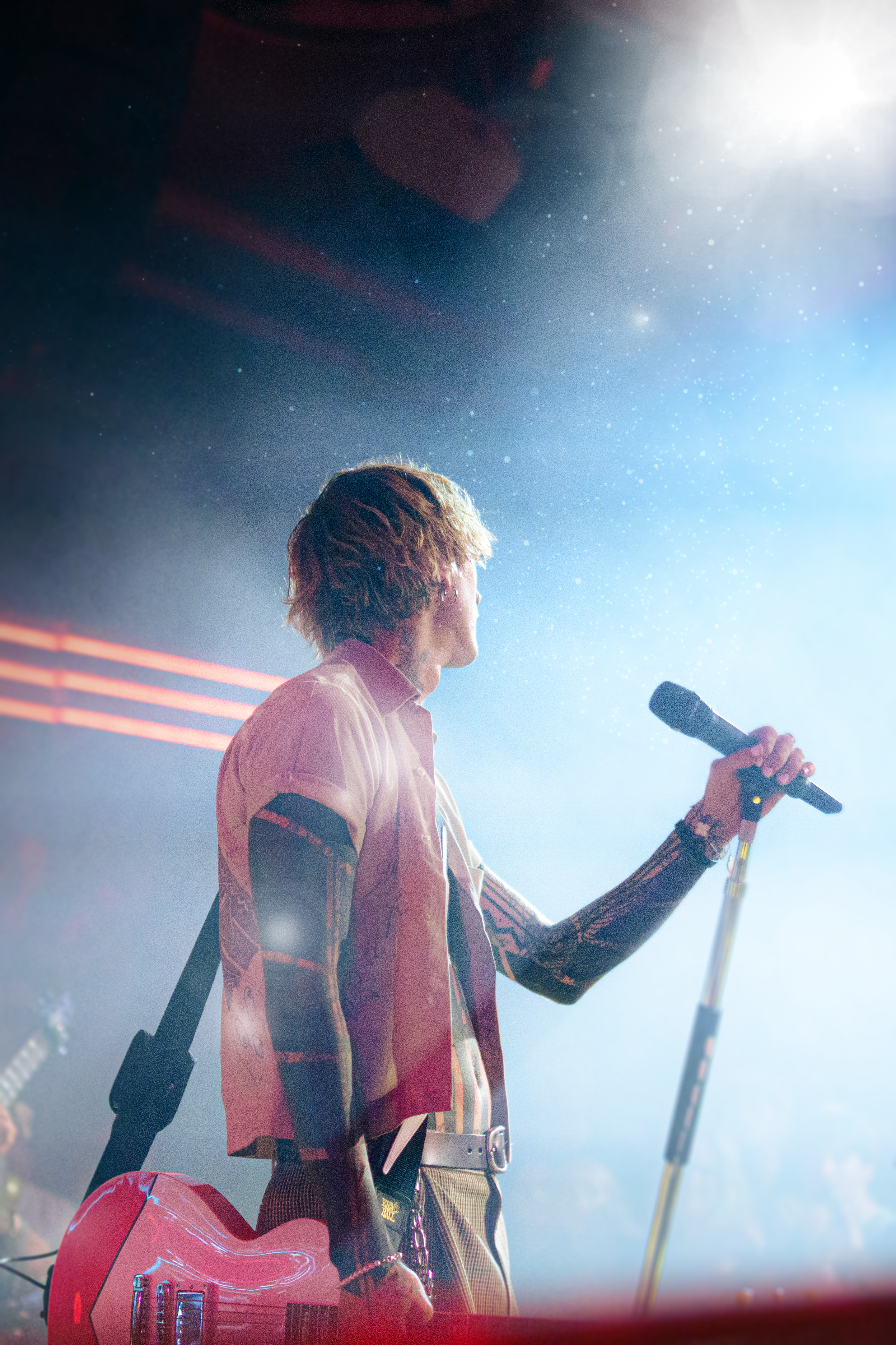 A male musician on stage holding a microphone, with a guitar hung over his shoulder, illuminated by stage lighting and looking towards the right.