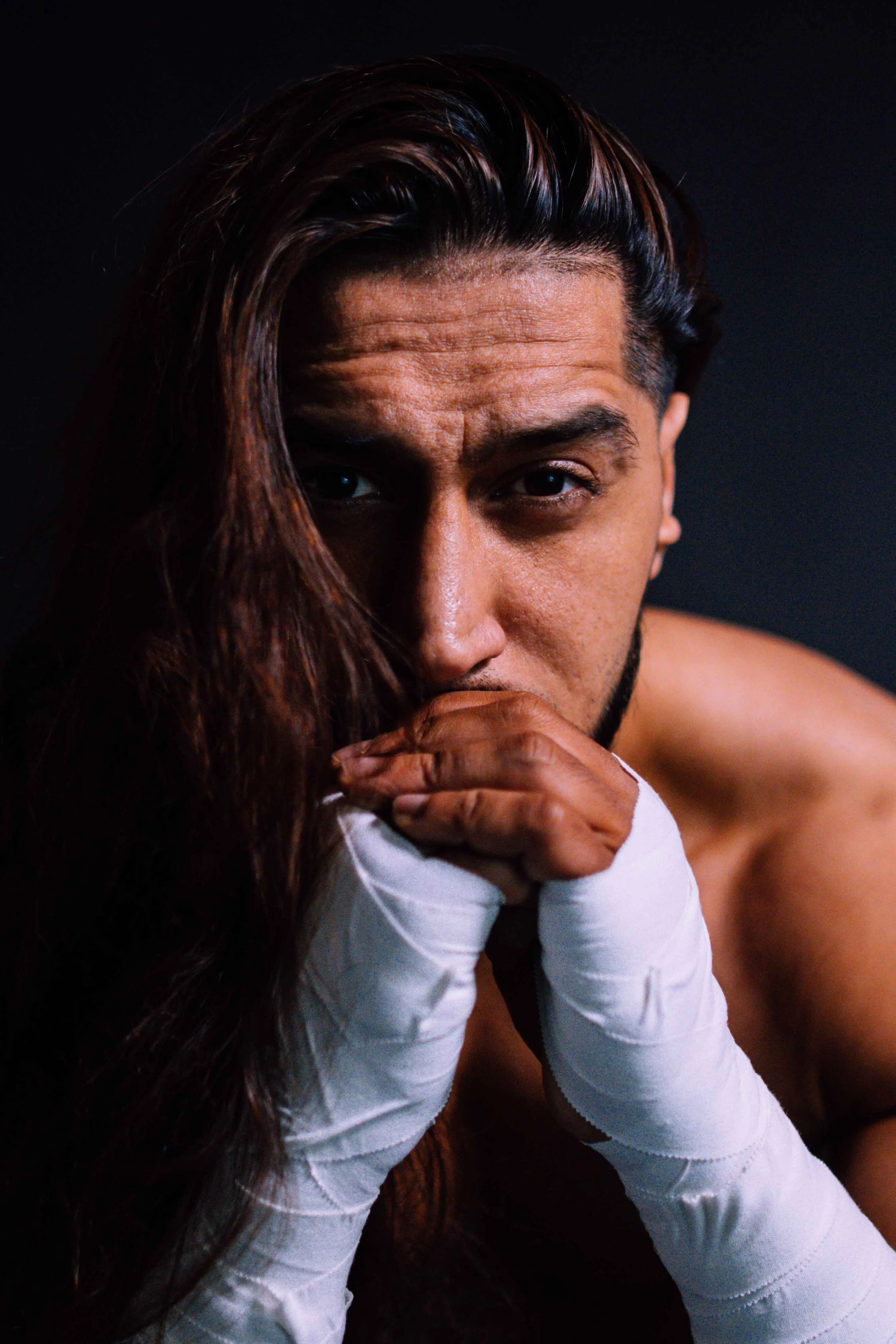 A shirtless man with dark hair styled to one side, resting his chin on his hands which are wrapped in white boxing wraps, looking directly at the camera against a black background.