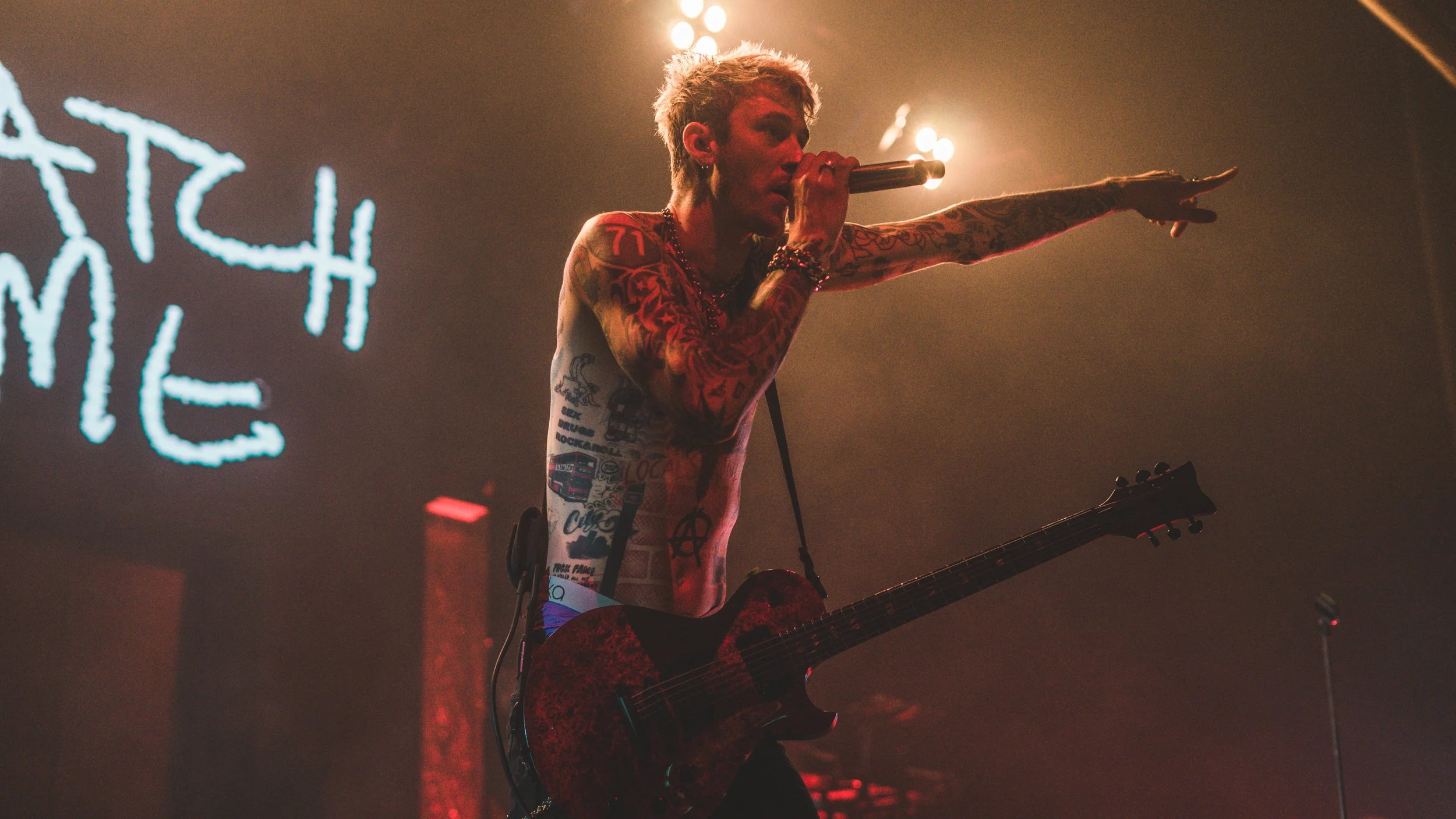 A tattooed male musician performing on stage with a guitar, holding a microphone, and pointing forward. Stage lights and a partially visible neon sign are in the background.