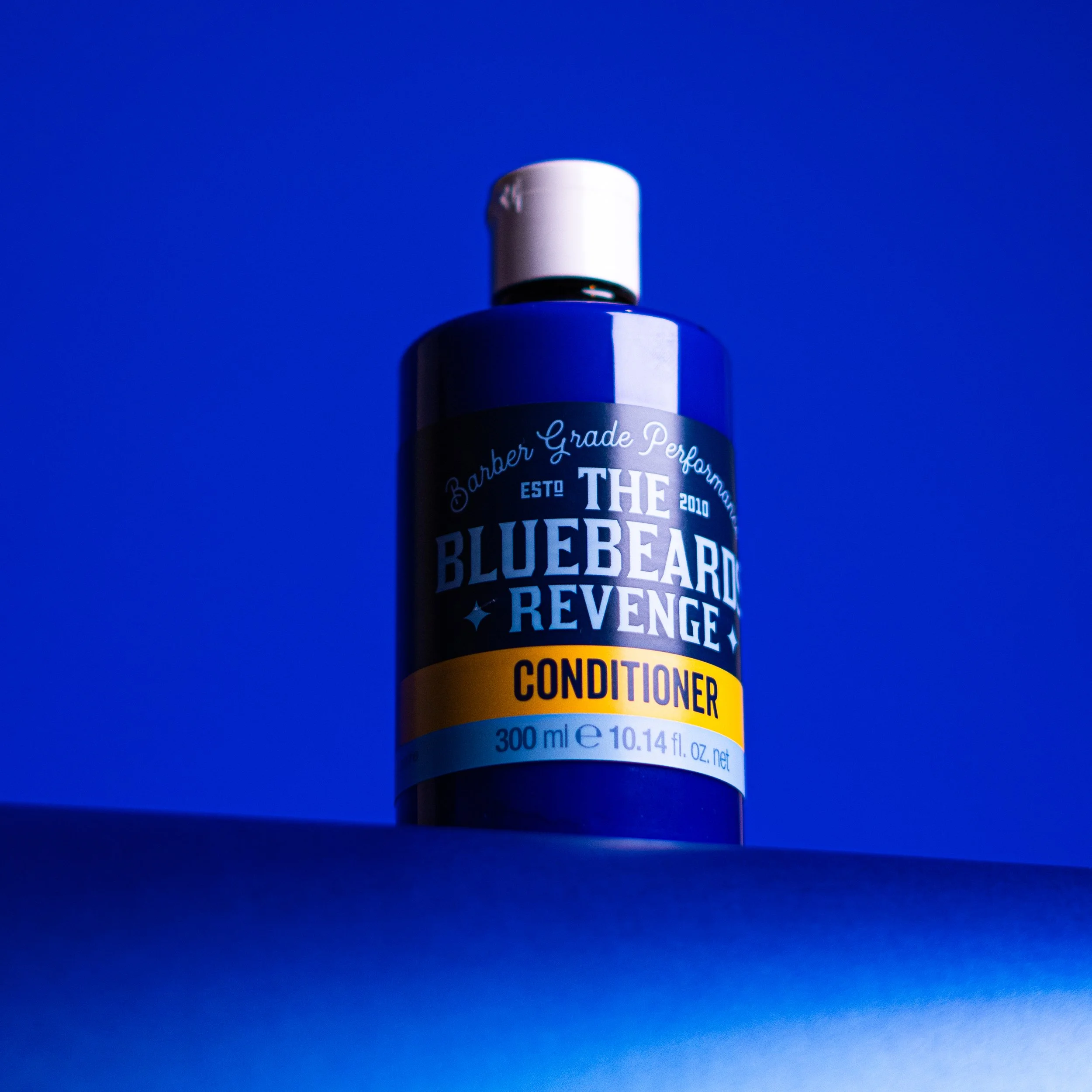 A bottle of Bluebeard Revenge conditioner with a dark blue body, white cap, and yellow label, placed on a surface against a vibrant blue background.